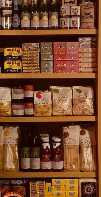 Shelf stocked with various food products including cans of sardines, jars of spreads, packets of pasta, and small boxes of snacks.