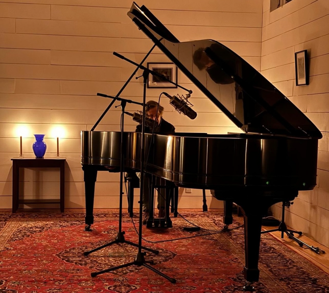 David Ellis Rissenberg playing on his 1895 Steinway concert grand piano in a cozy, warmly lit studio with wooden walls, a table with a blue vase and candles, and framed photos on the wall.