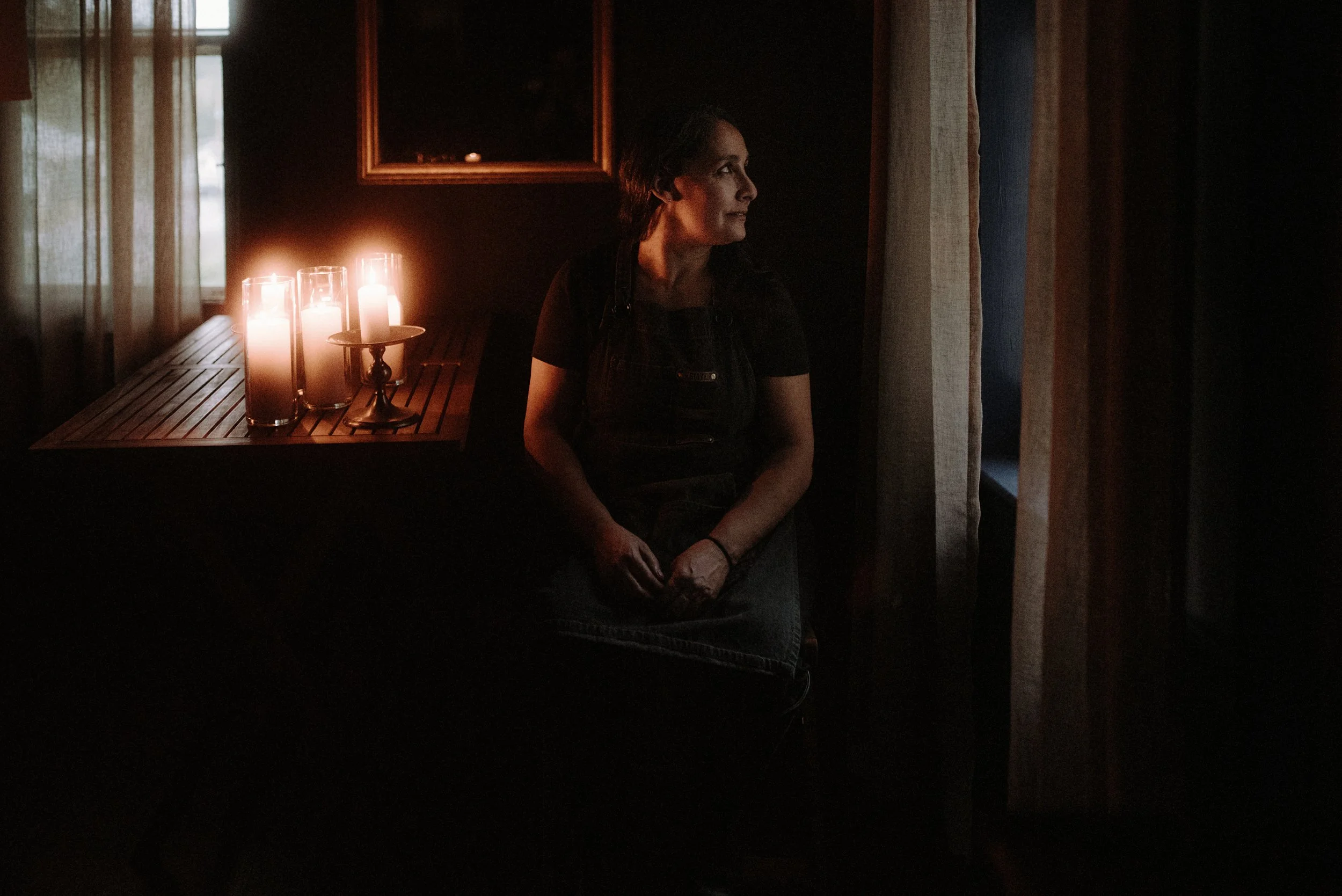A woman sitting in a dark room illuminated by candlelight, looking out a window, wearing a black shirt and apron.