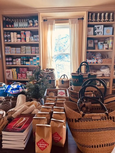 A cozy store display with coffee bags, books, woven baskets, and shelves stocked with various canned and packaged goods, with a window in the back bringing in natural light.