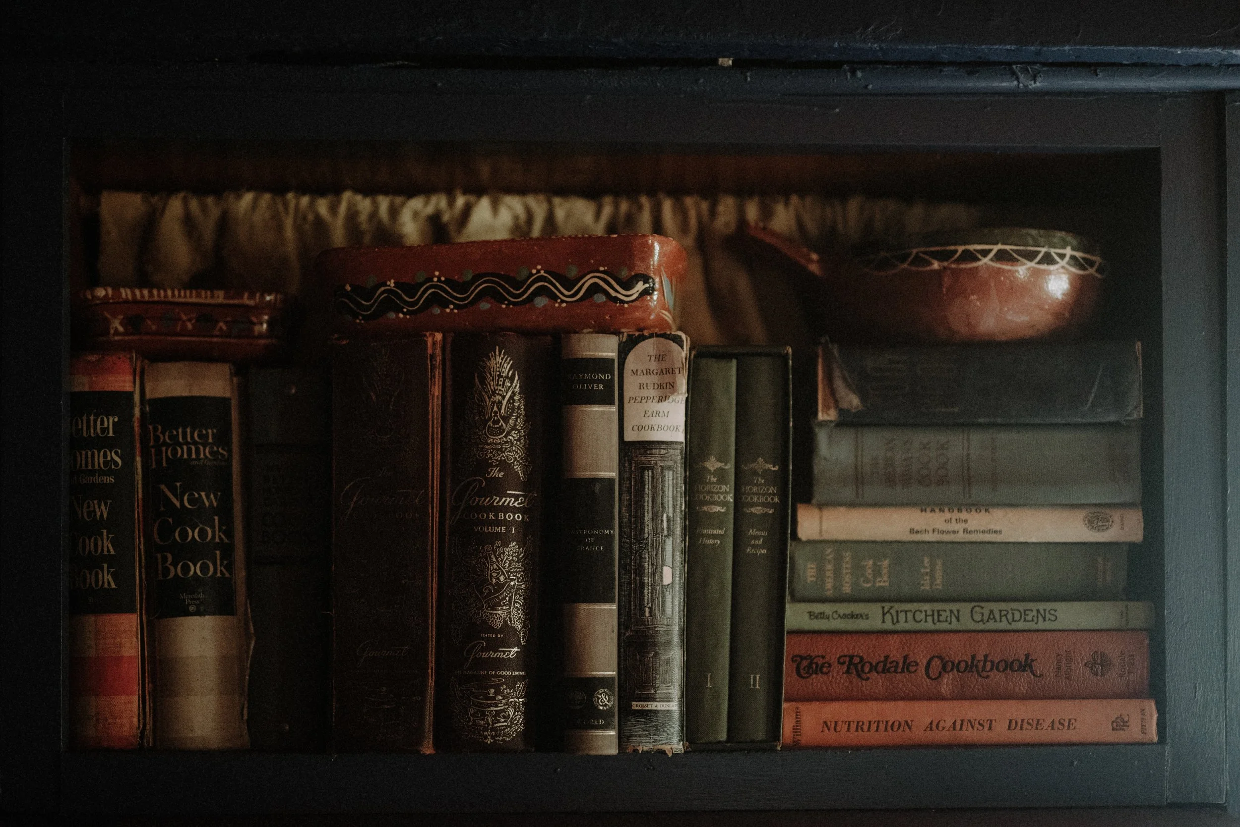 A dark wooden shelf containing a variety of old, vintage cookbooks and decorative tins. The titles include 'Better Homes and Gardens New Cookbook,' 'The Gourmet Cookbook,' 'The Horizon Cookbook,' and several others, with some tins on top of the books and a fabric curtain in the background.