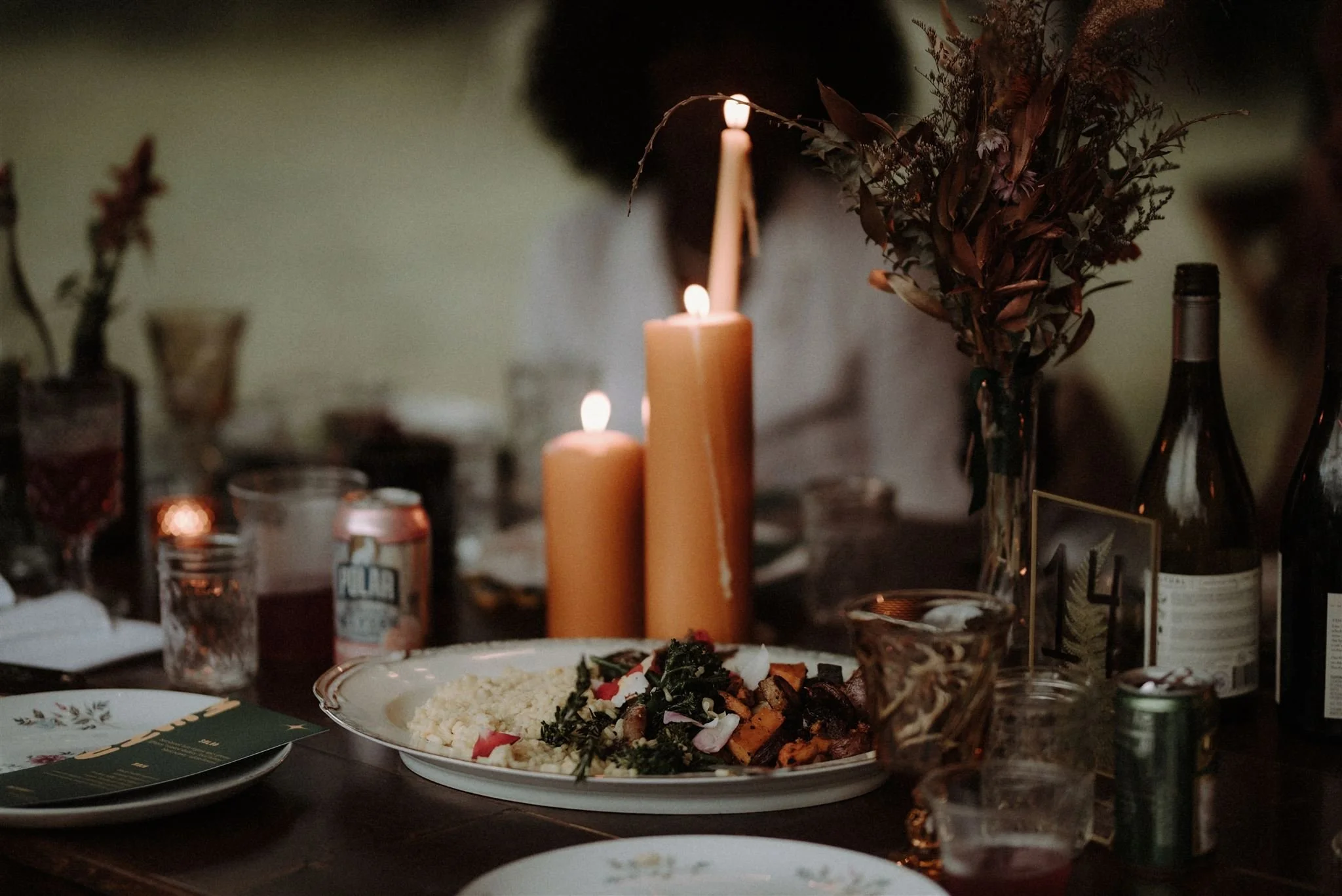 A dinner table with candles, a plate of food, beverages, and a floral centerpiece.