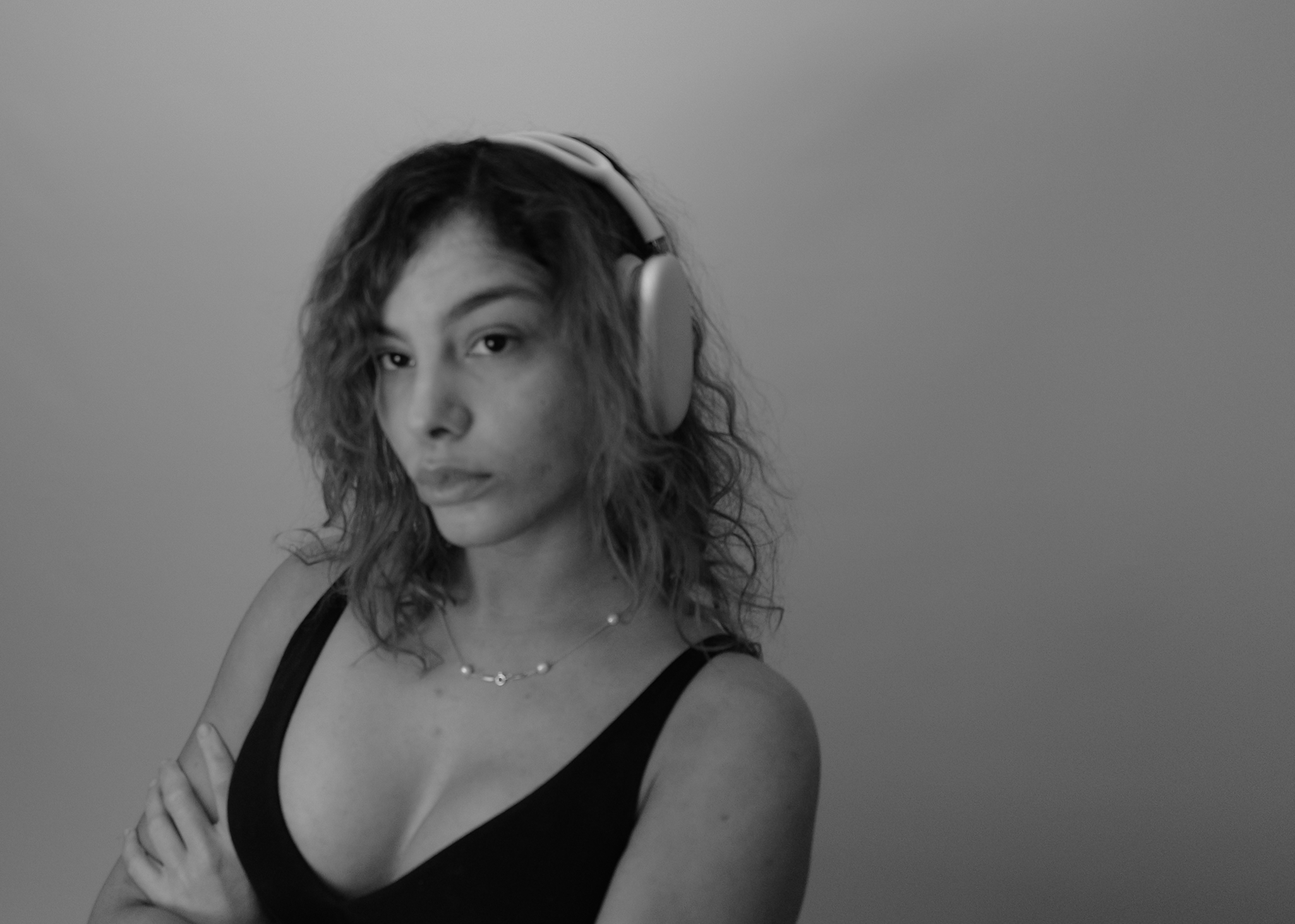 A woman with curly hair wearing headphones, a black top, and a necklace, looking at the camera with a serious expression, standing against a plain background.