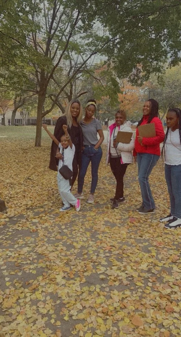 Group of five women and one young girl standing outdoors on a leaf-covered ground with trees in the background, enjoying fall weather.