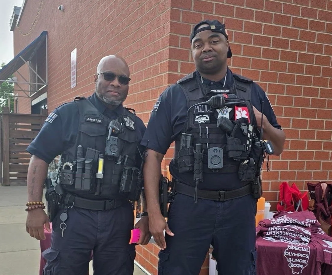 Two police officers standing outdoors in front of a brick wall. They are wearing tactical uniforms with various equipment and badges. One officer is wearing sunglasses and the other a backwards cap. To the right, there is a table with maroon shirts and other items.