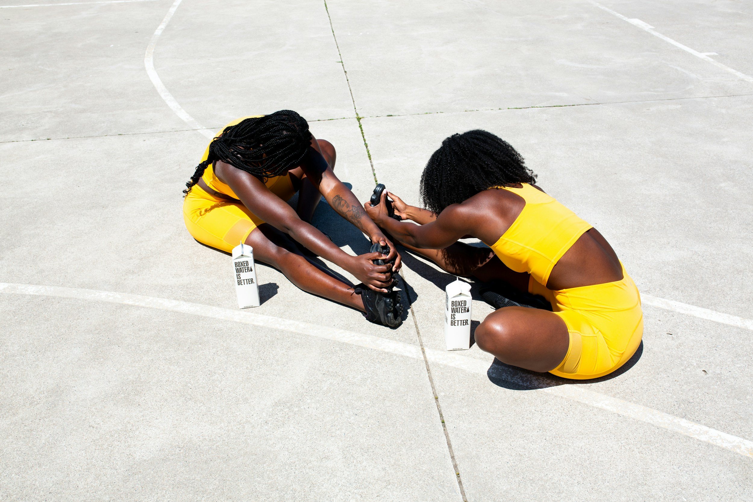 Two women in yellow athletic wear stretching on a concrete sports court, sitting on the ground and holding their feet with their hands, with water bottles nearby that read 'Boxed water is better'.