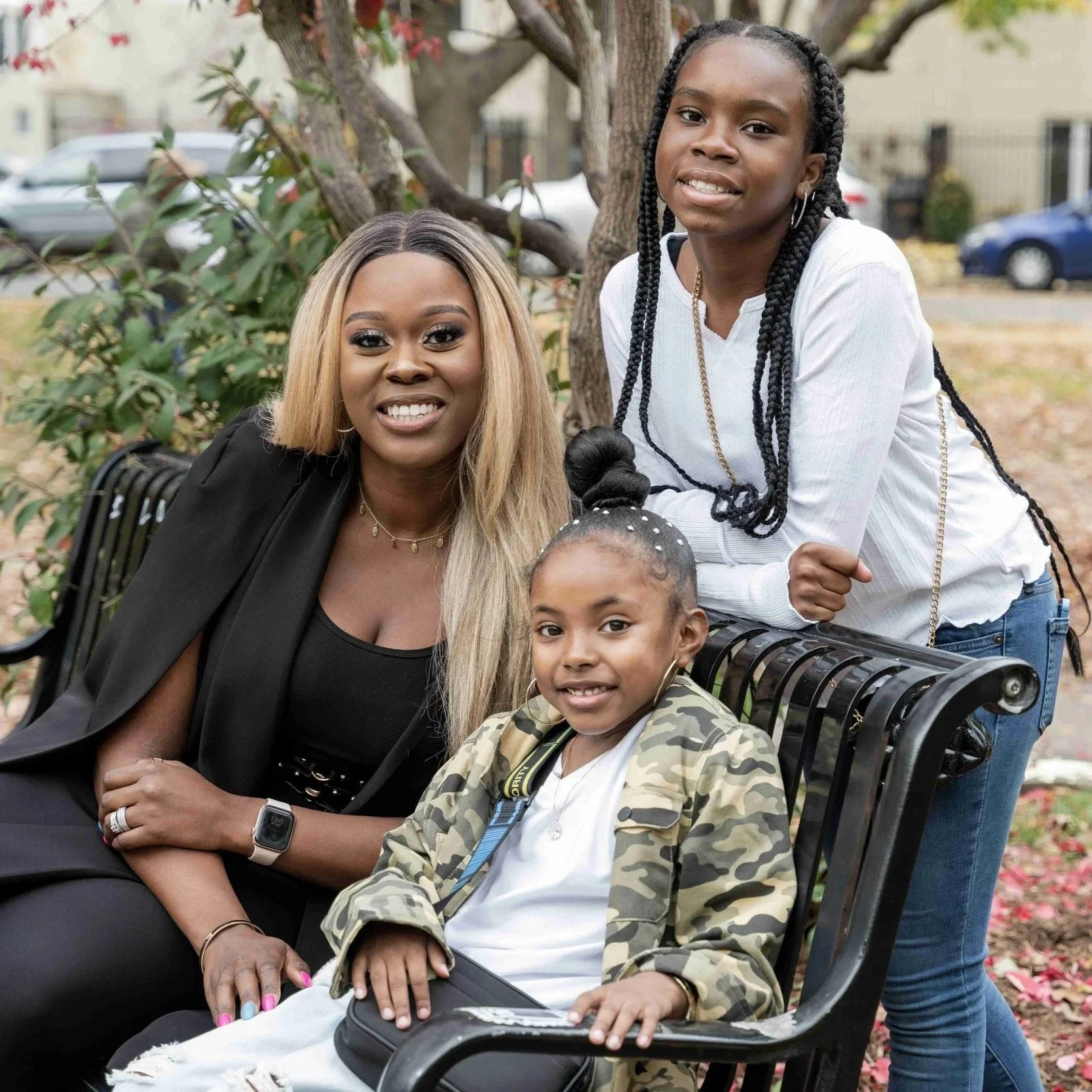 A woman and two young girls are outdoors sitting and standing near a park bench in a yard with a tree and parked cars in the background. The woman has long blonde hair, and the girl standing has long braided hair. The girl sitting has her hair in a bun with decorative pearls, and is wearing a camouflage jacket. All three are smiling.