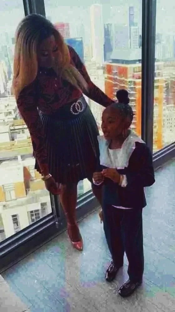 A woman leaning over to touch or check the hair of a young girl, both standing near a large window with a city skyline in the background. The girl is smiling and looking up at the woman.