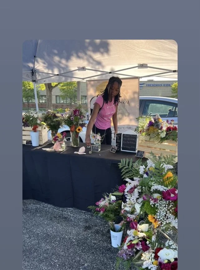 A woman arranging flowers at a flower booth under a white canopy. The booth displays various flower arrangements and small chalkboard signs. There are more flowers on the ground and in buckets, with a background of trees, a building, and a parked car.