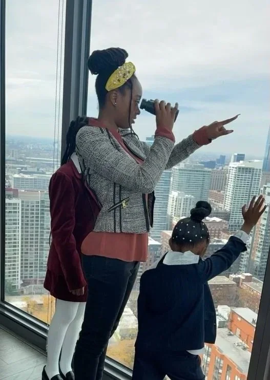 A woman and two children looking out of a high-rise building window at the city skyline. The woman is holding binoculars and pointing, while one child is pointing out the window.