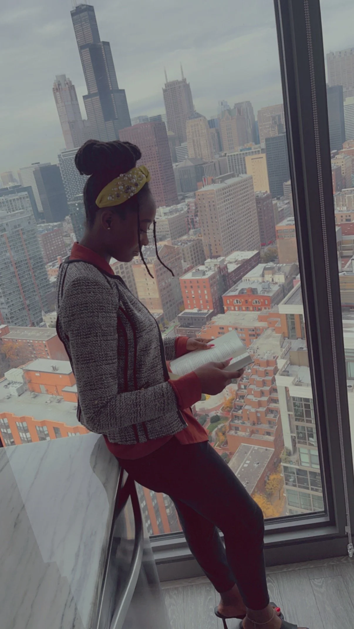A young woman with braided hair and a yellow headband walking and reading a book on a high balcony in a city with tall buildings.