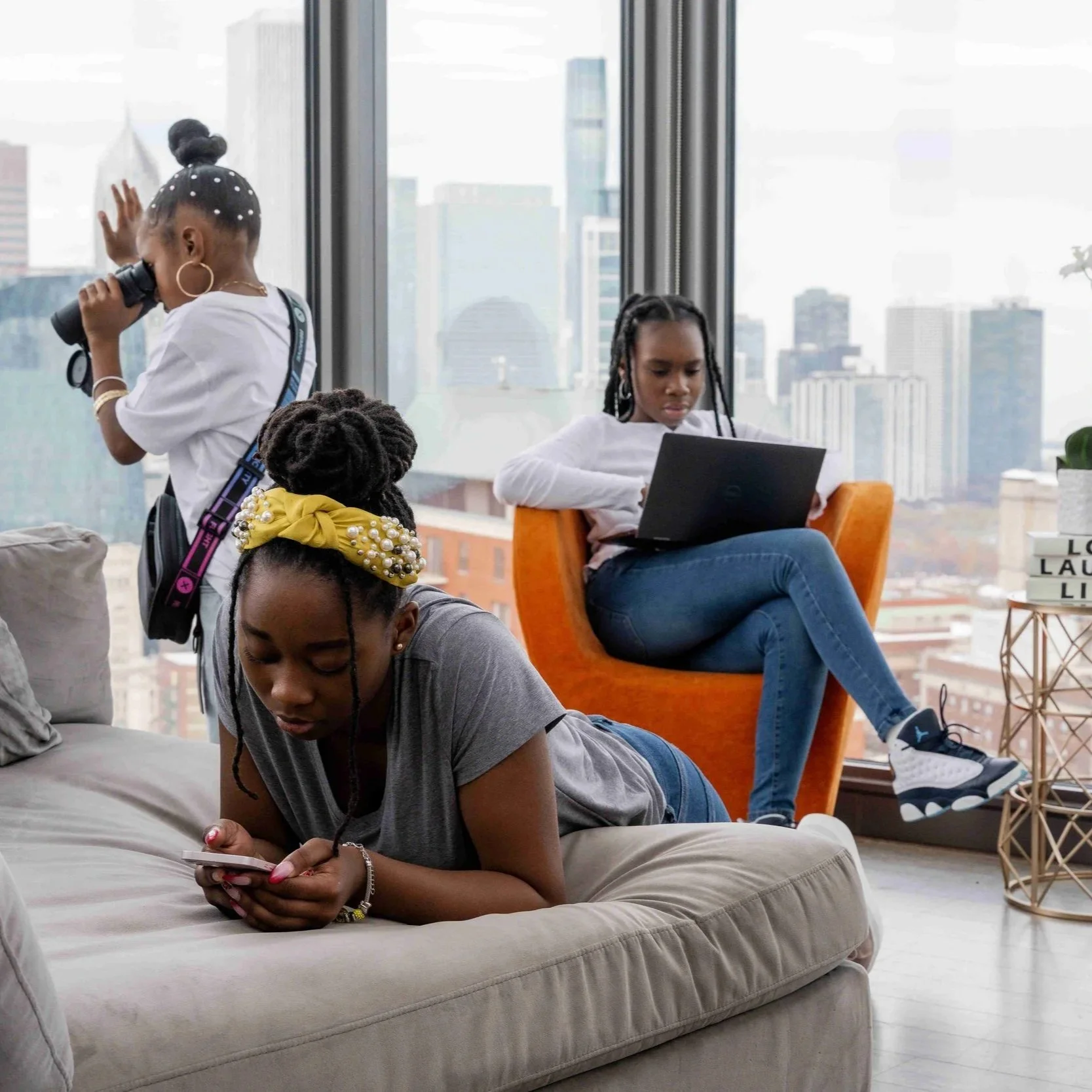 Four young girl friends spending time indoors in a city apartment with large windows showing city skyscrapers. One girl is lying on the sofa using her phone, another sitting in an orange chair working on a laptop, a third girl standing and looking through binoculars, and the fourth girl is on the floor looking at her phone.