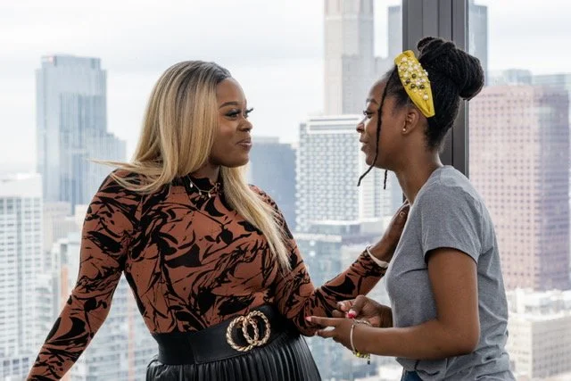 A woman leaning over to touch or check the hair of a young girl, both standing near a large window with a city skyline in the background. The girl is smiling and looking up at the woman.