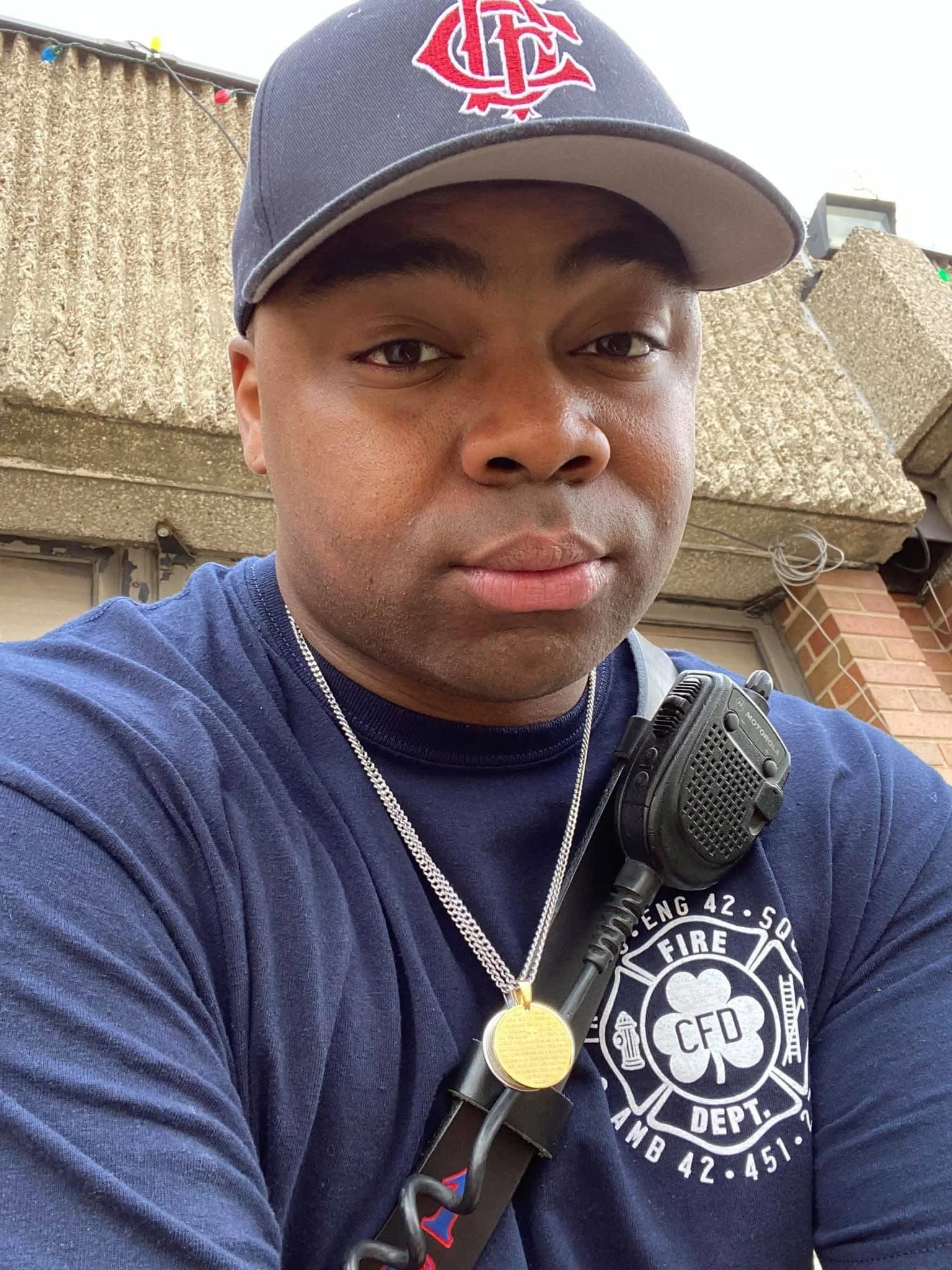 A firefighter wearing a navy blue shirt with fire department insignia, a necklace with a gold medallion, a radio on his shoulder, and a baseball cap with a red logo.
