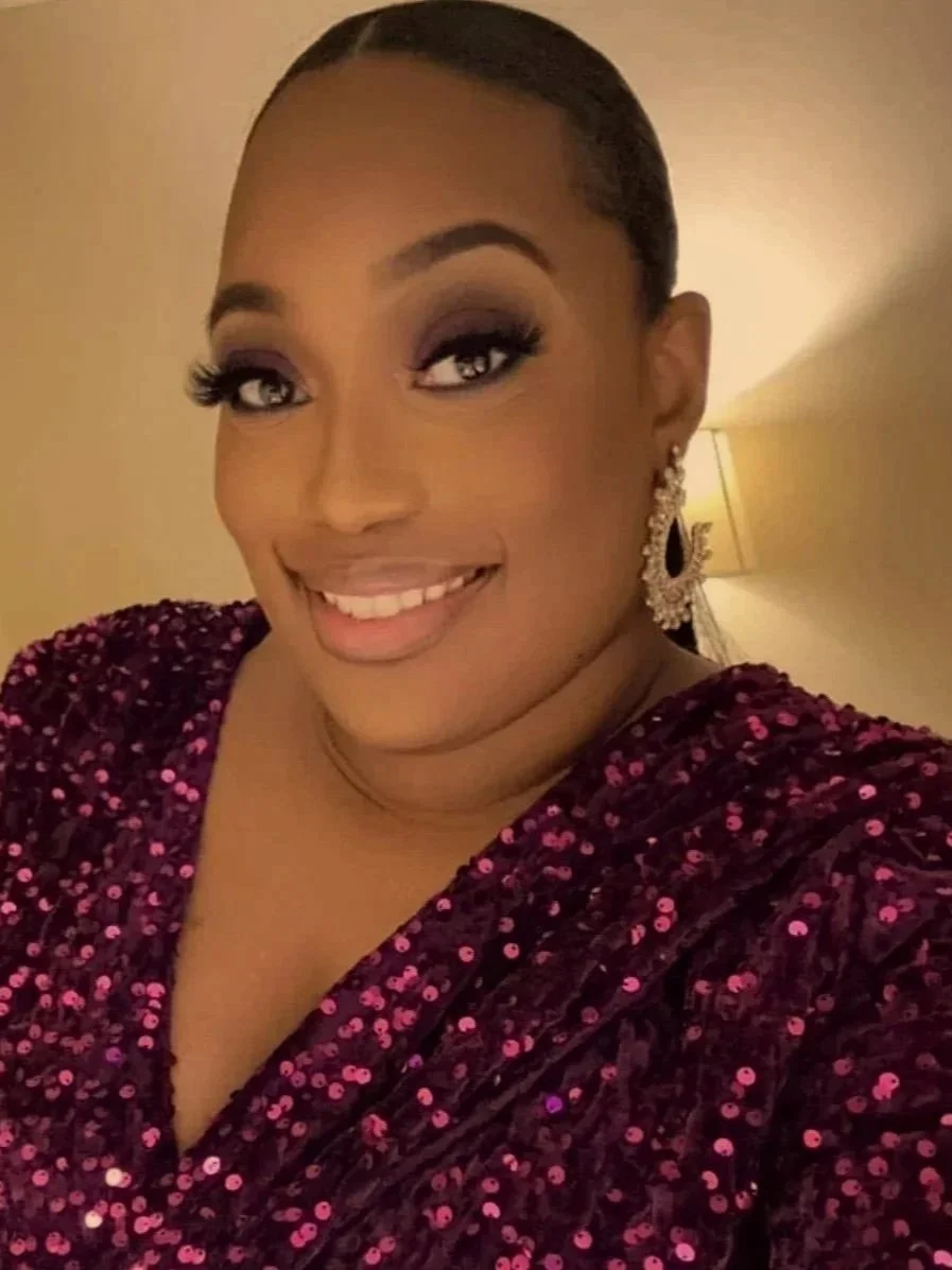 A woman smiling, wearing a dark purple sequin dress and large earrings, with makeup and her hair styled back. She is in a warmly lit room.