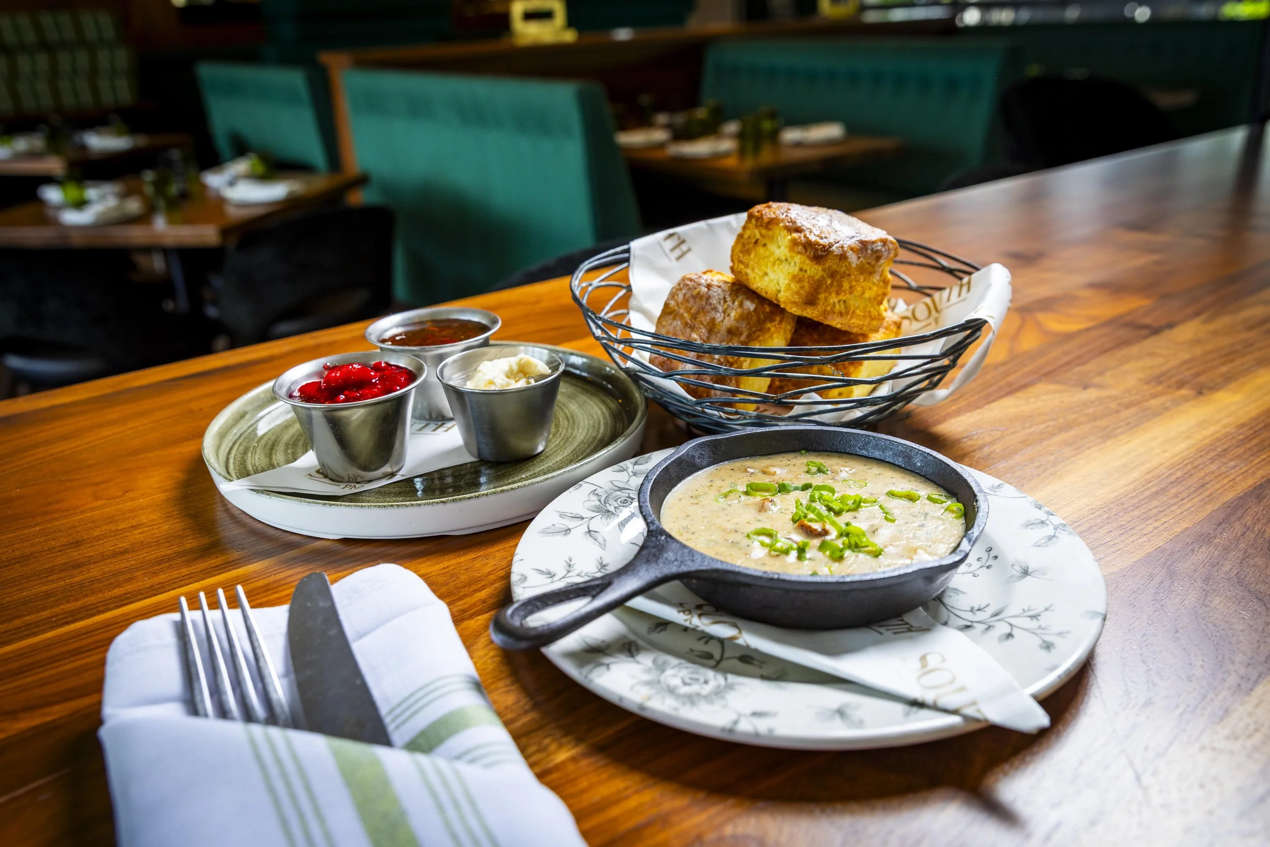 A table with a serving of creamy soup garnished with green onions in a cast iron skillet, a basket of golden biscuits, and small metal cups containing whipped cream, cranberry sauce, and gravy, set in a restaurant with green booths in the background.