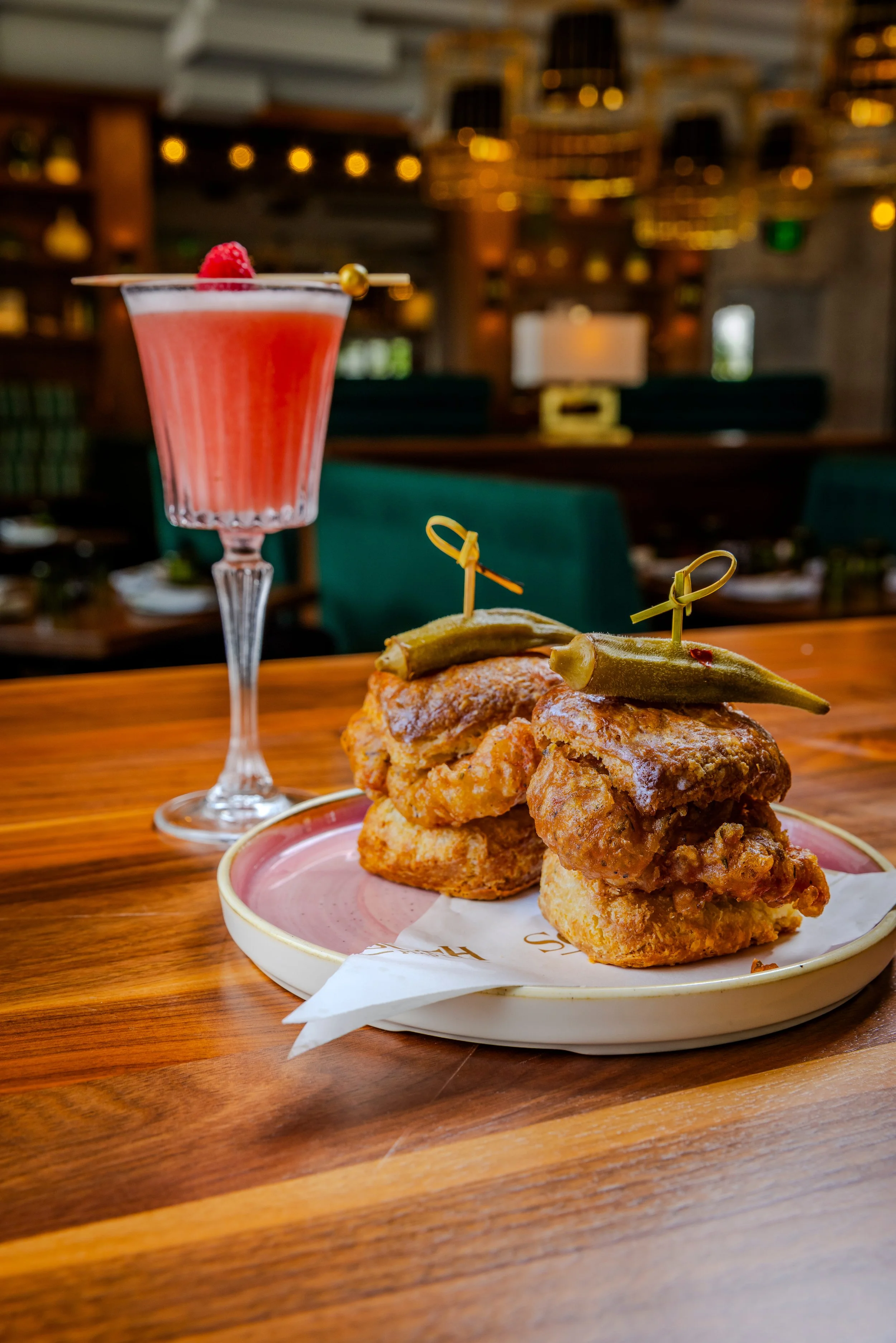 Red cocktail beside fried chicken biscuits topped with pickles on a wooden bar.