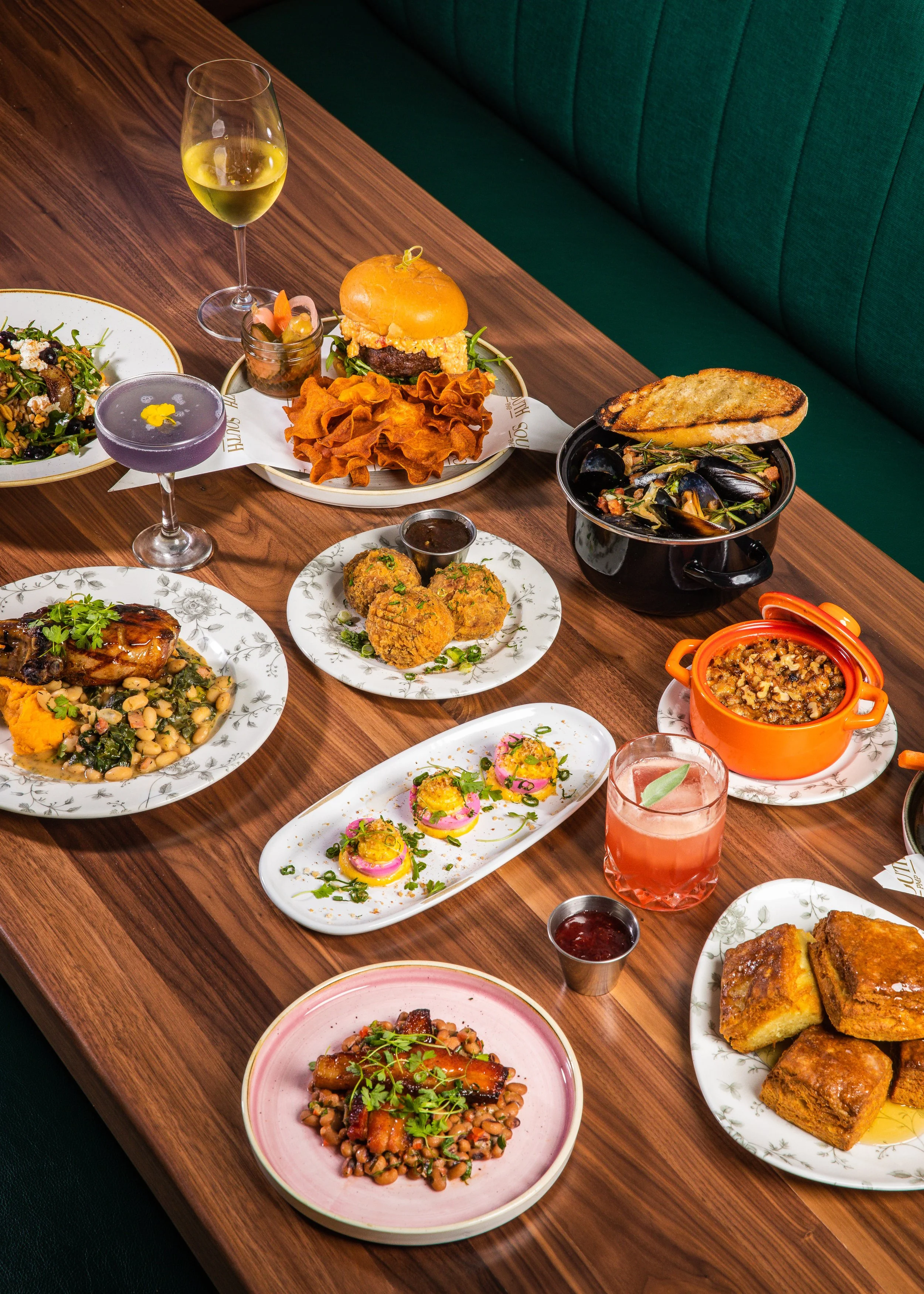 A table with a variety of dishes including a burger, seafood, salad, and desserts, along with drinks like wine and cocktails, set for a meal.