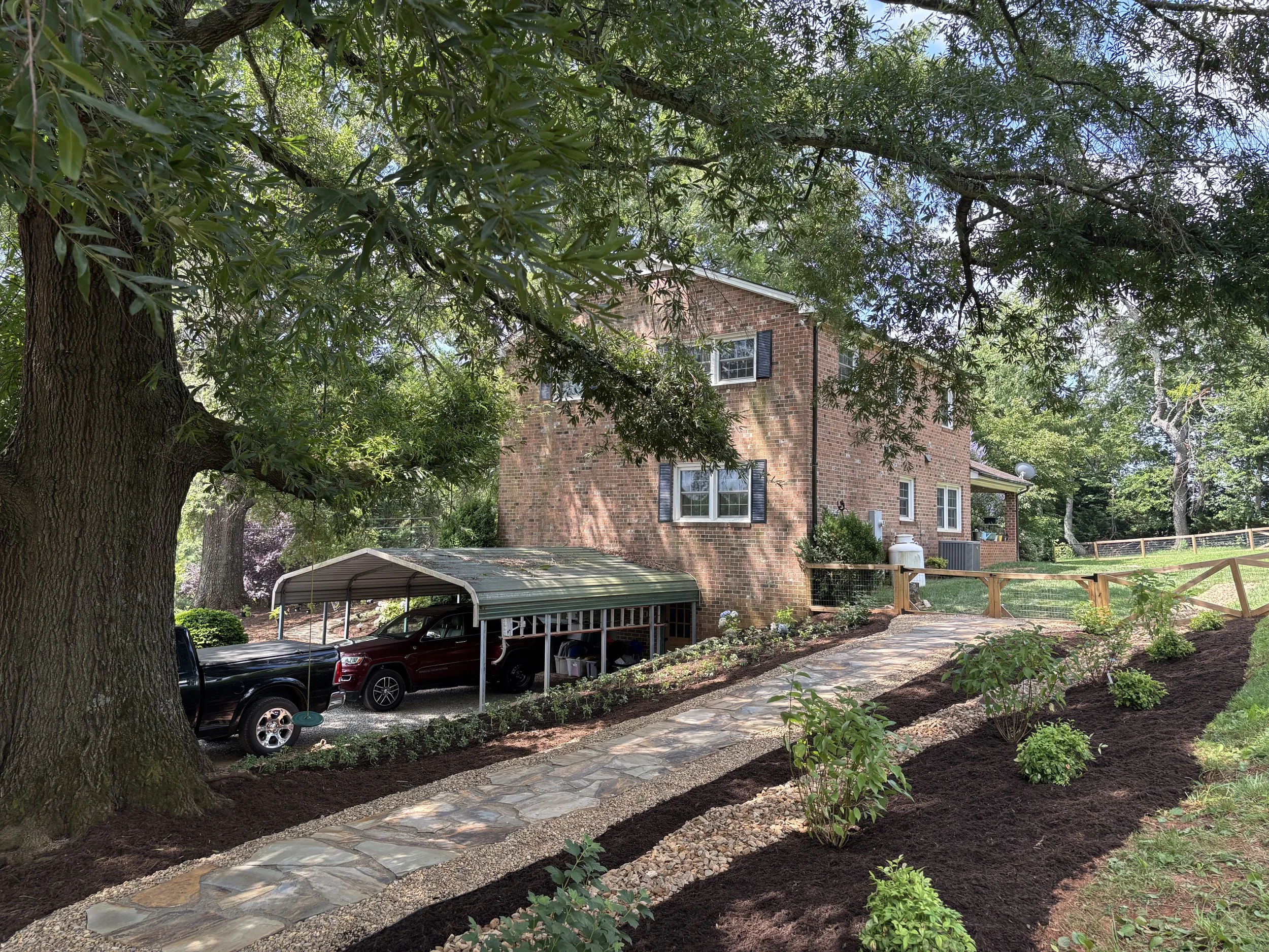 A landscaped backyard with a stone pathway, trimmed shrubs, large trees, a brick house with black shutters, a covered parking area for vehicles, and a wooden fence.