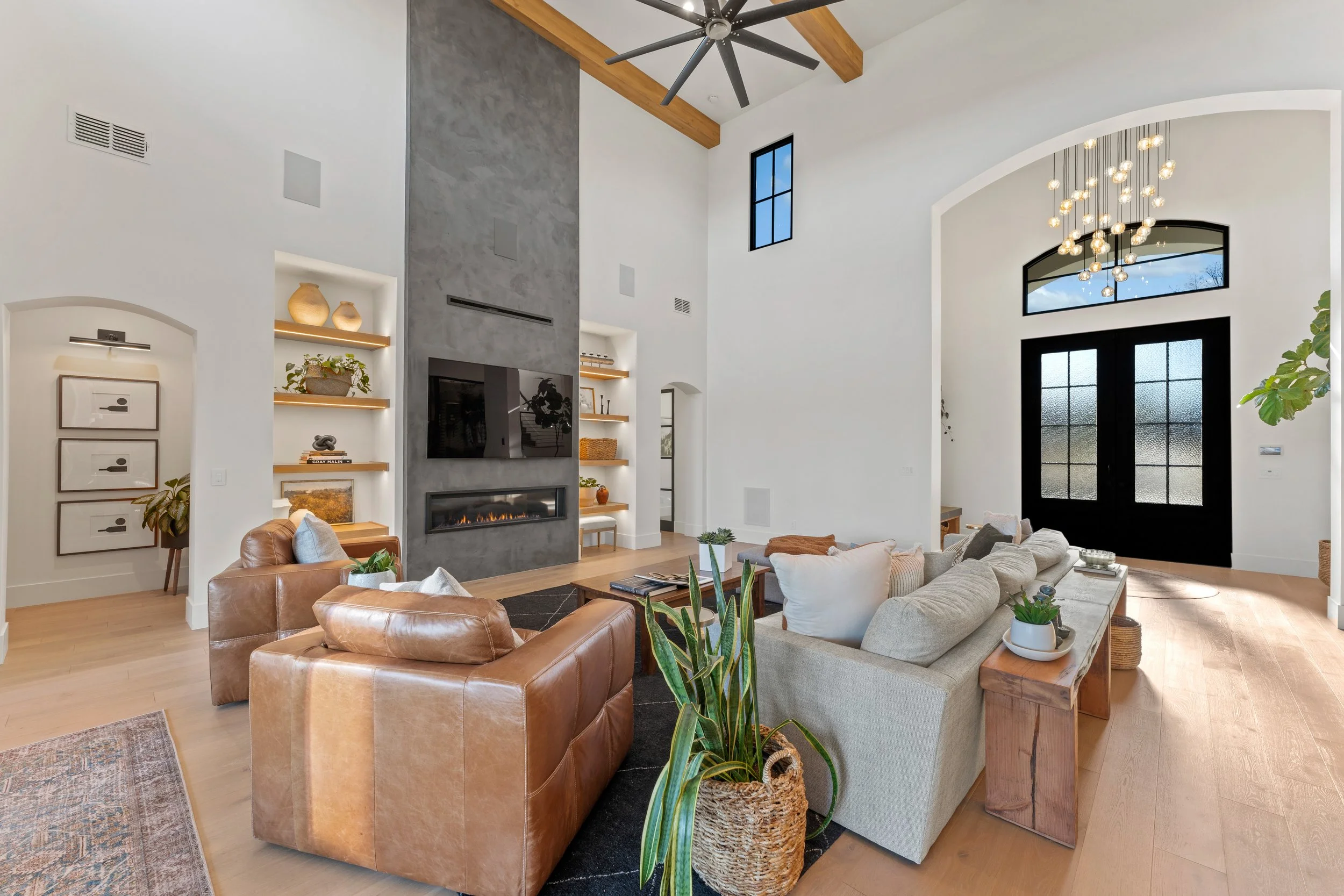 Spacious living room with high ceiling, large black front door with glass panels, and a modern chandelier. Features a fireplace, a large beige sectional sofa, leather armchair, wooden accents, and built-in shelves with decorative items.