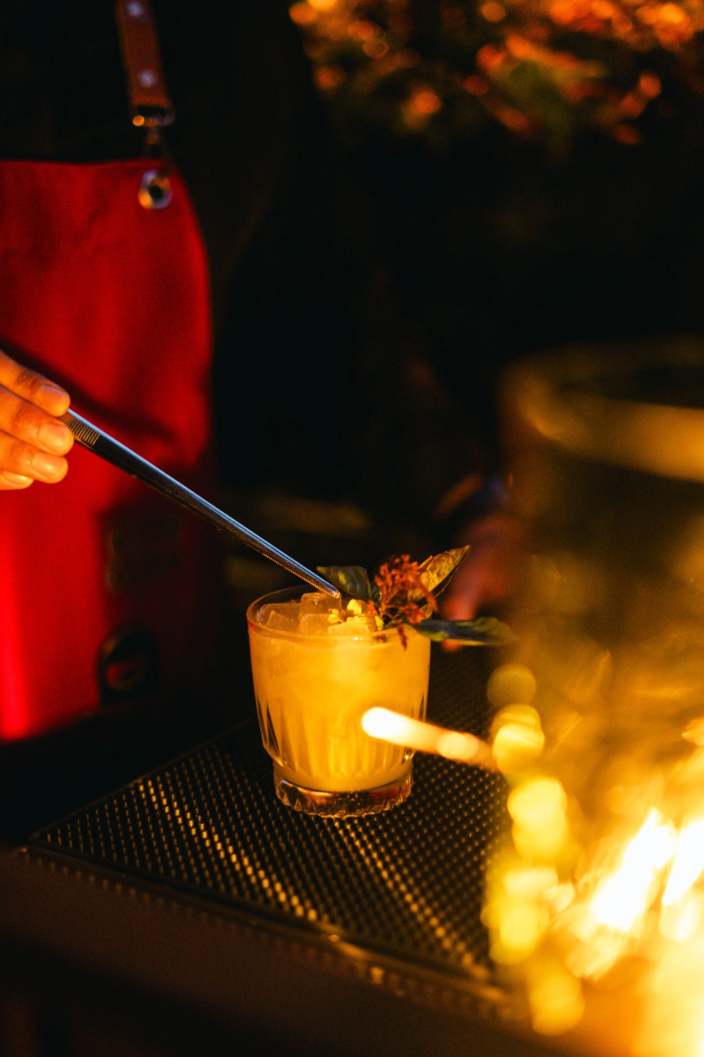 A bartender adding finishing touches to a yellow cocktail with herbs and ice in a glass, illuminated by warm, low lighting.