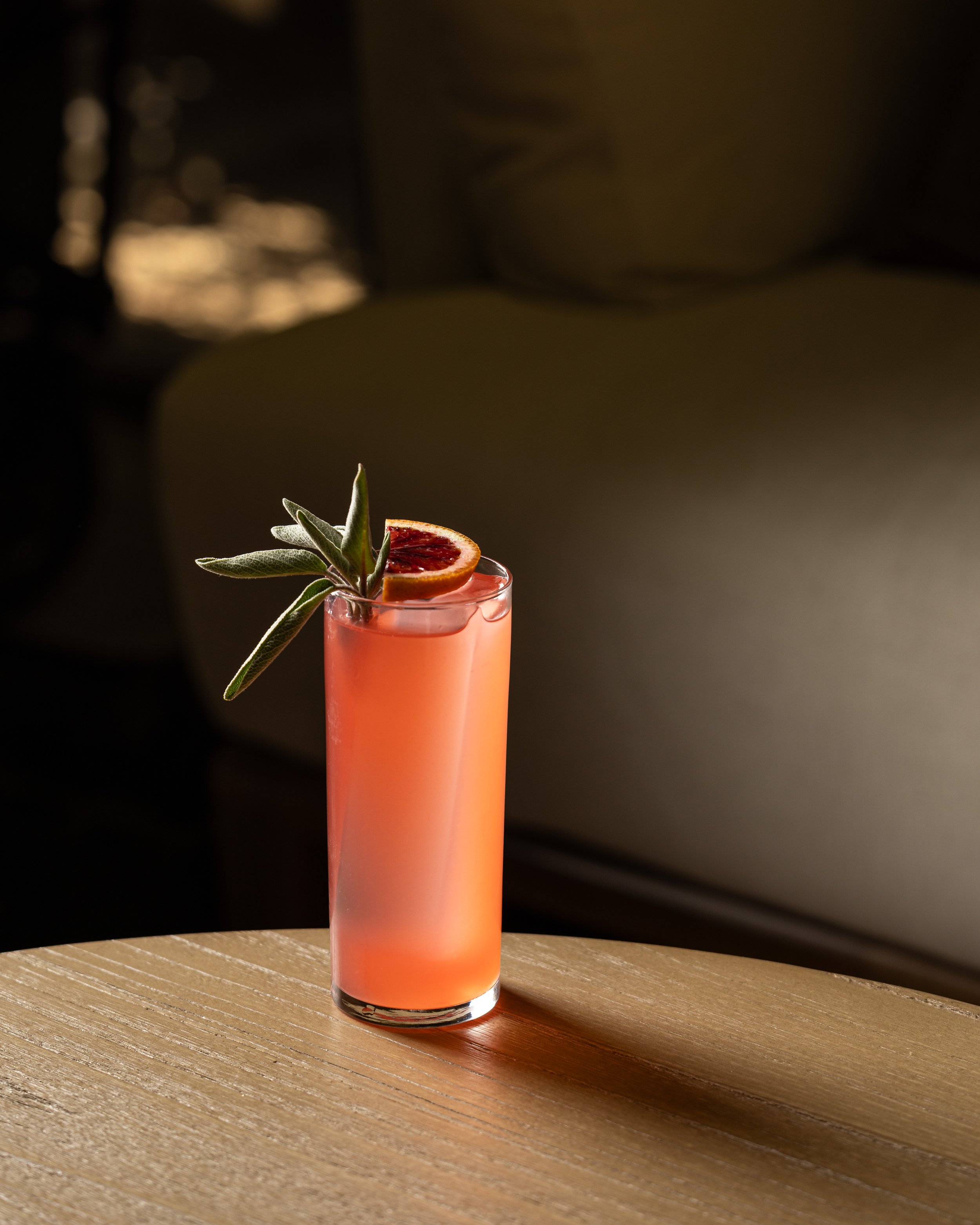 A tall glass of pink cocktail garnished with a dried blood orange slice and sage leaves on a wooden surface.