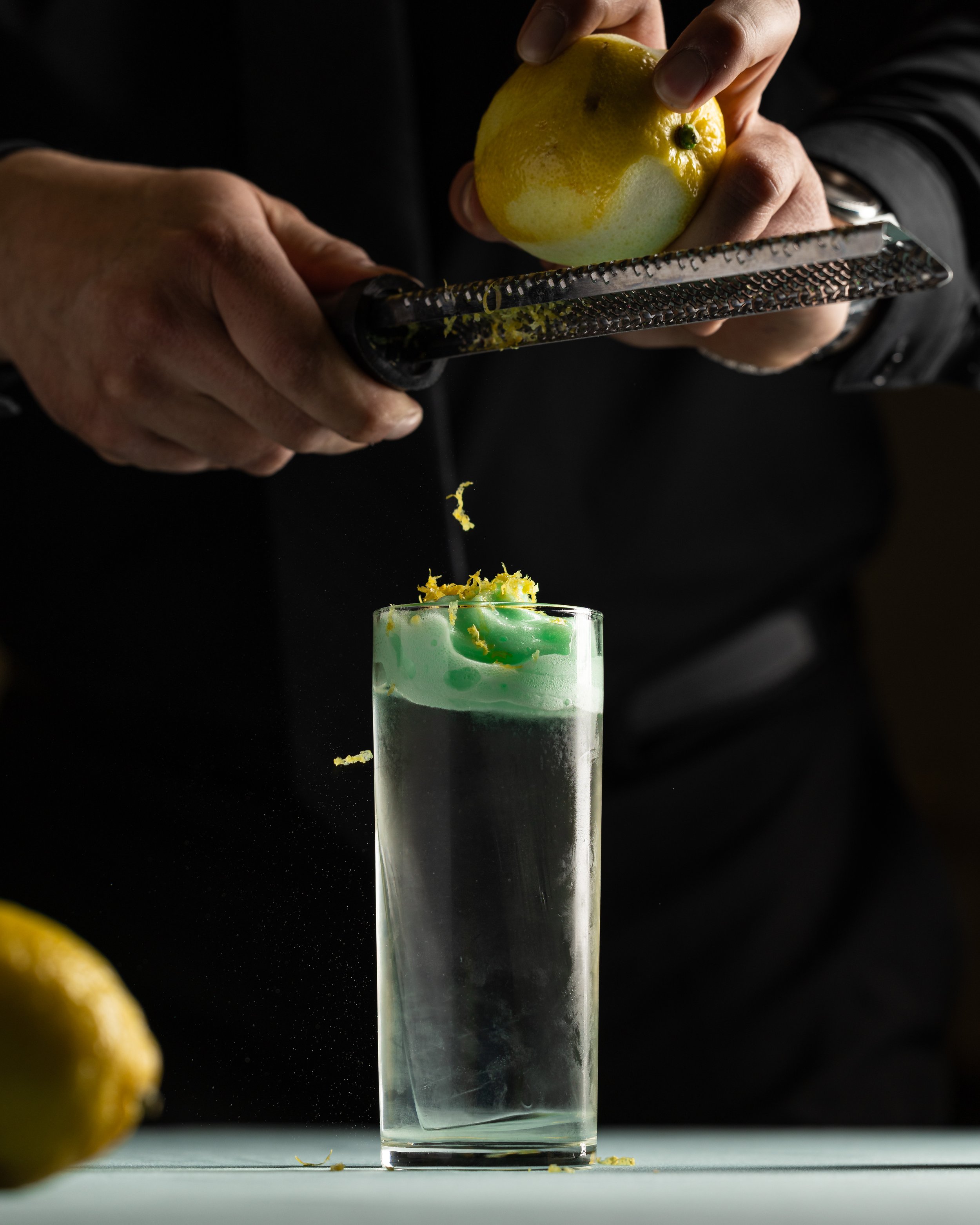 A person grating lemon peel over a cocktail glass filled with a green drink, with lemon peel falling into the glass, on a white surface.