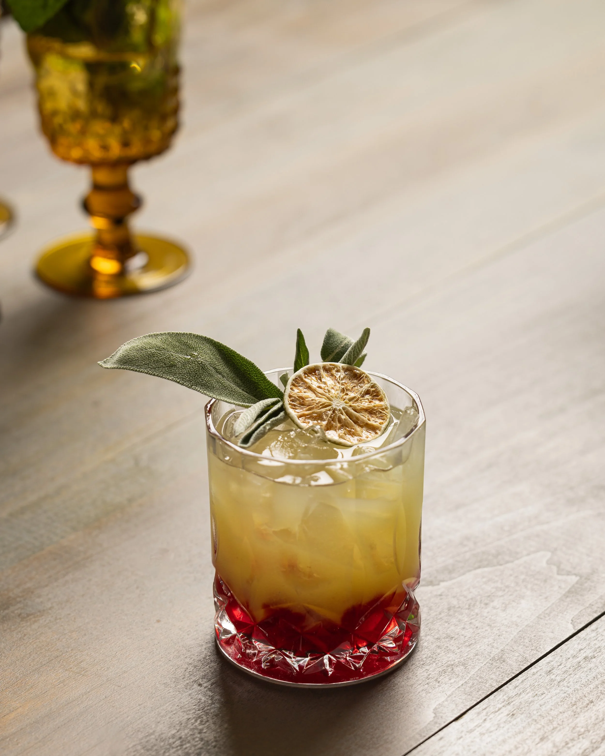 Colorful cocktail with ice, garnished with a dried citrus slice and sage leaves in a patterned glass on a wooden surface.