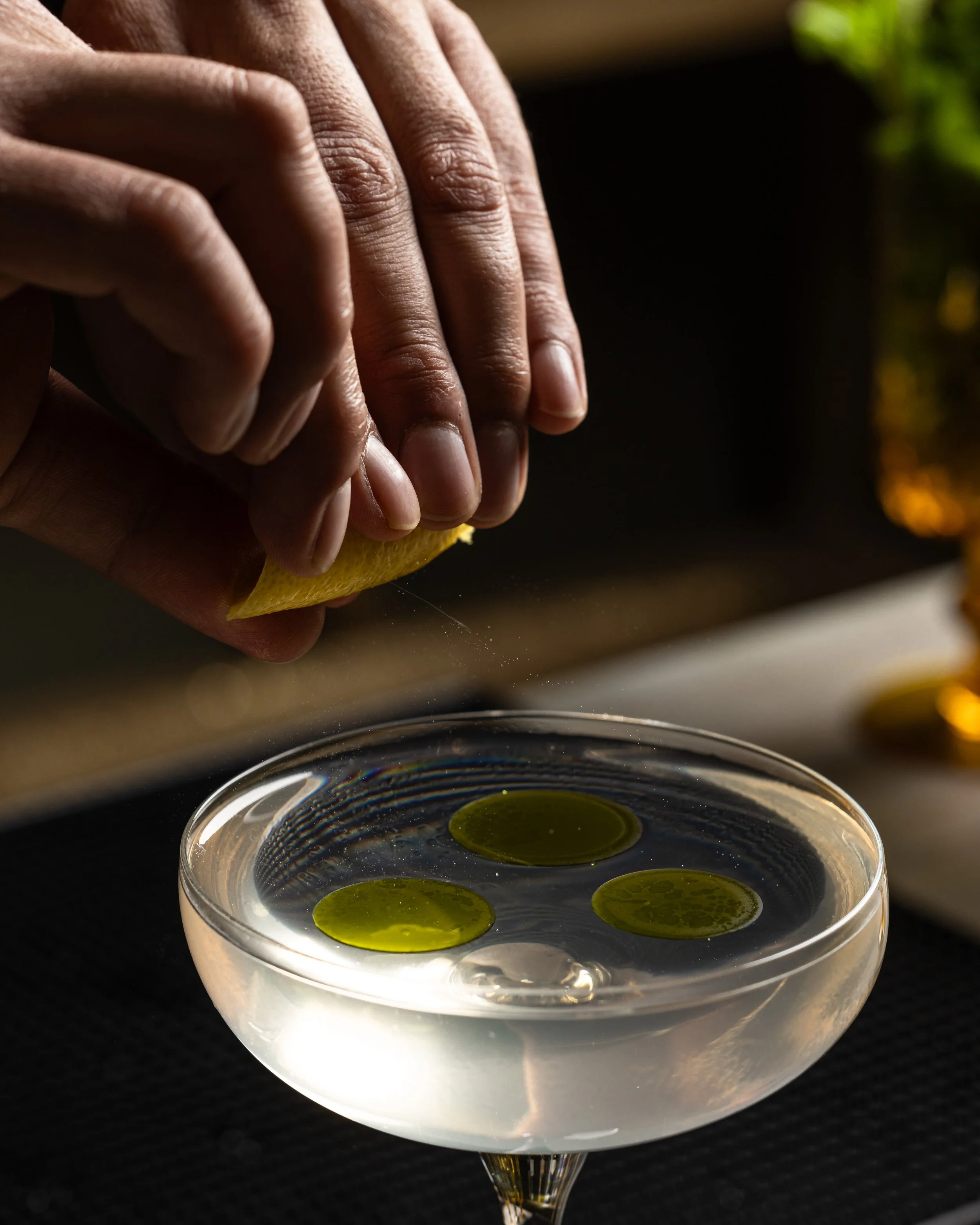 Person squeezing lemon peel over a cocktail glass with three green olive slices in liquid.