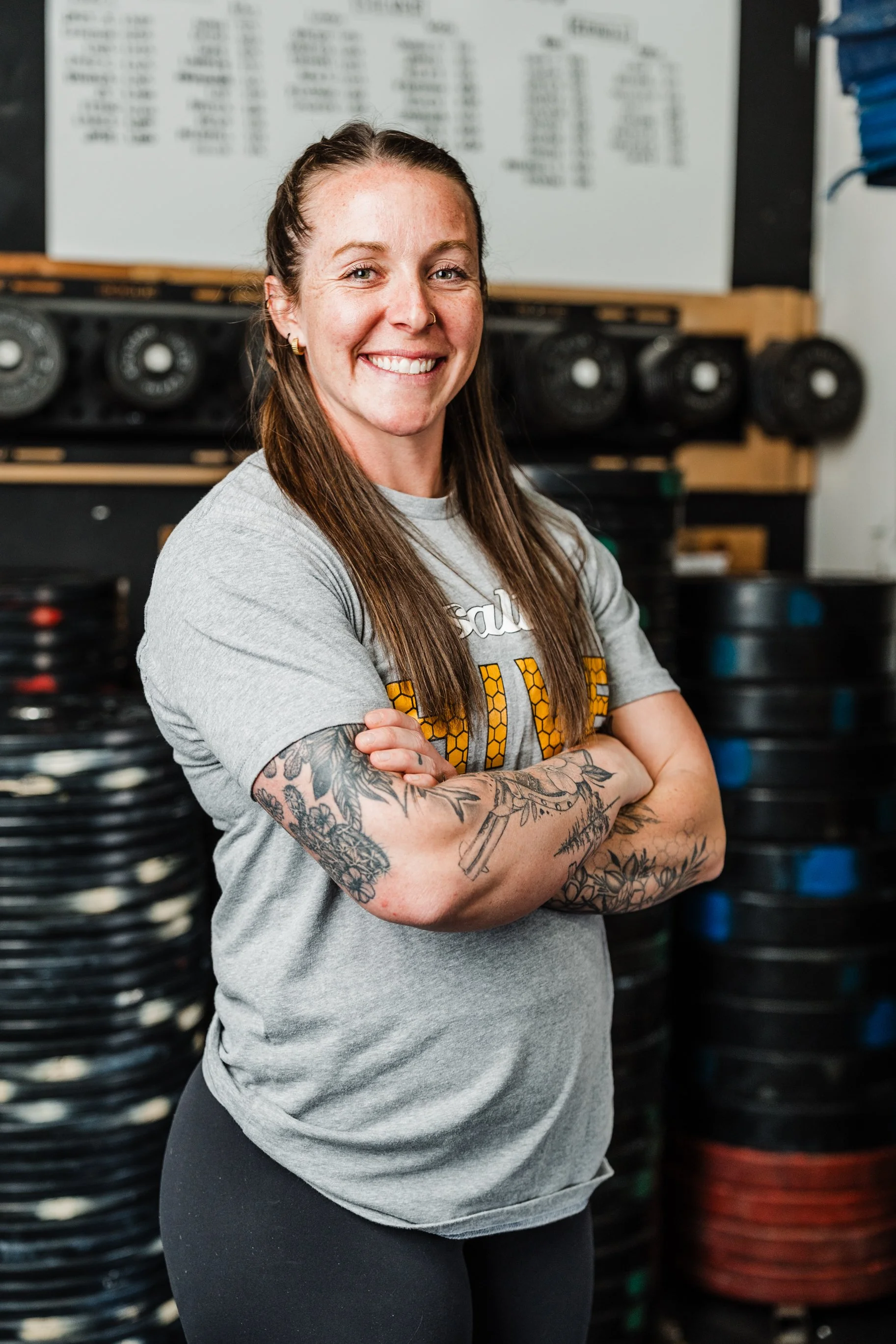 A smiling woman with long brown hair, tattoos on her arms, and a nose ring, standing in a gym with black weight plates and a whiteboard in the background.