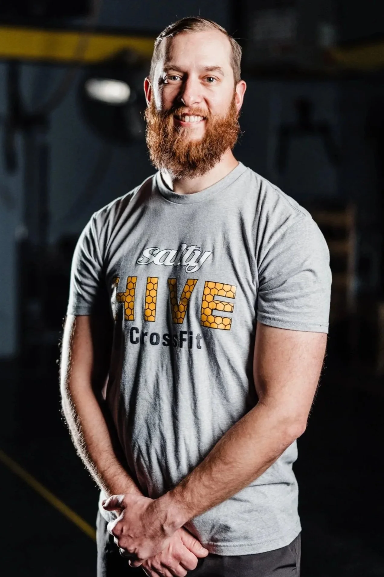A man with a red beard smiling, wearing a gray T-shirt with the words 'salty HIVE CrossFit' printed on it, standing in a gym.