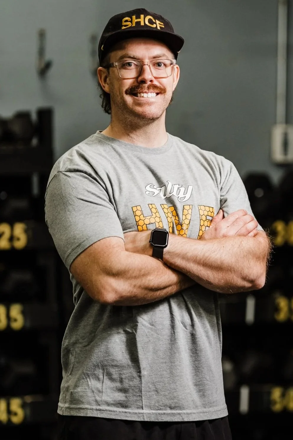 A man with glasses, a mustache, and a beard smiling with arms crossed, wearing a black cap with yellow letters 'SHCF,' a grey t-shirt, and a black smartwatch, standing in front of a background of black weight plates marked with yellow numbers.