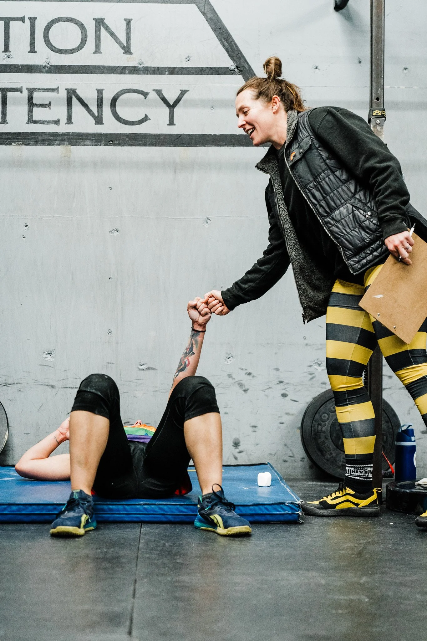 A woman in workout clothes, including yellow and black striped leggings, and a black jacket, is standing and smiling while helping a person lying on the floor during an exercise session, in front of a wall with a partially visible emergency sign.
