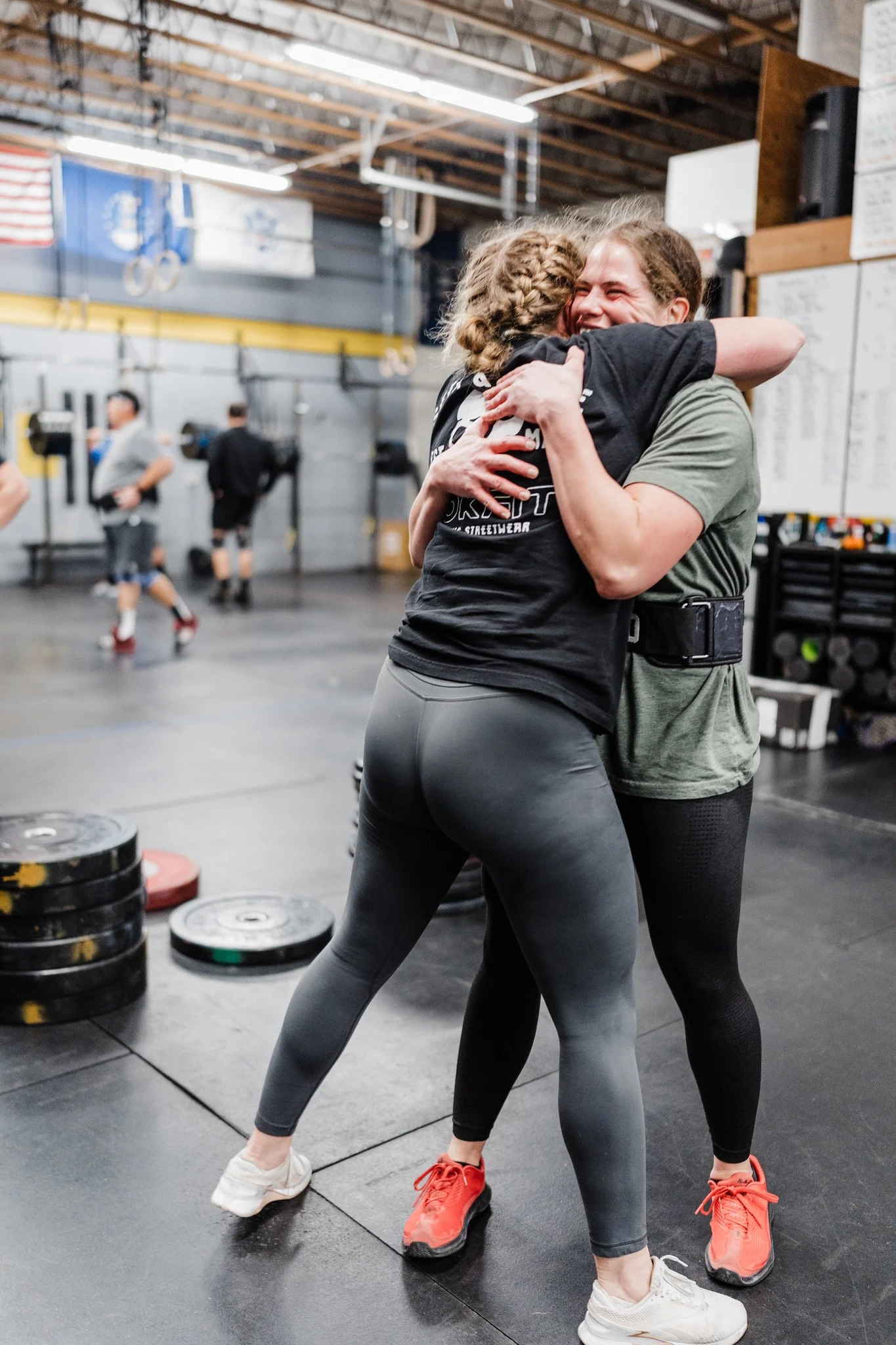 Two women hugs in a gym with weights and other people working out in the background.