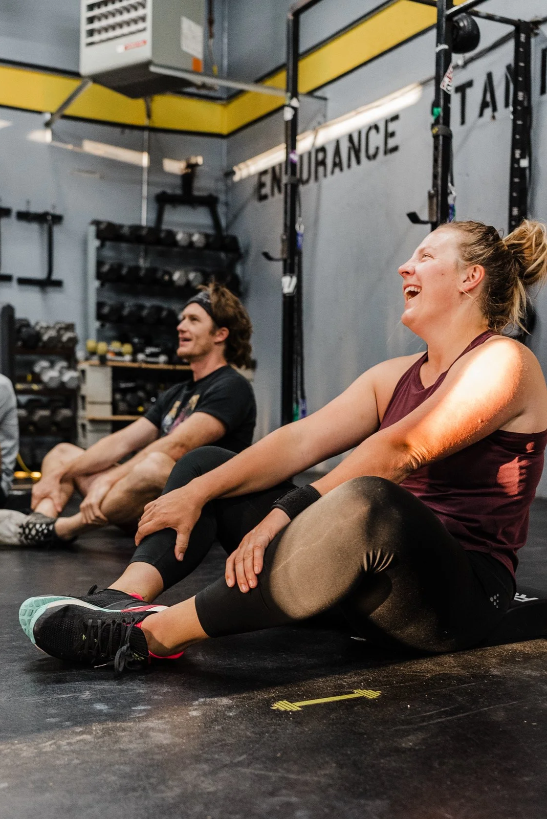 Two people sitting on the floor inside a gym, smiling and laughing after a workout session. The background shows gym equipment and a wall with the word 'Endurance'.