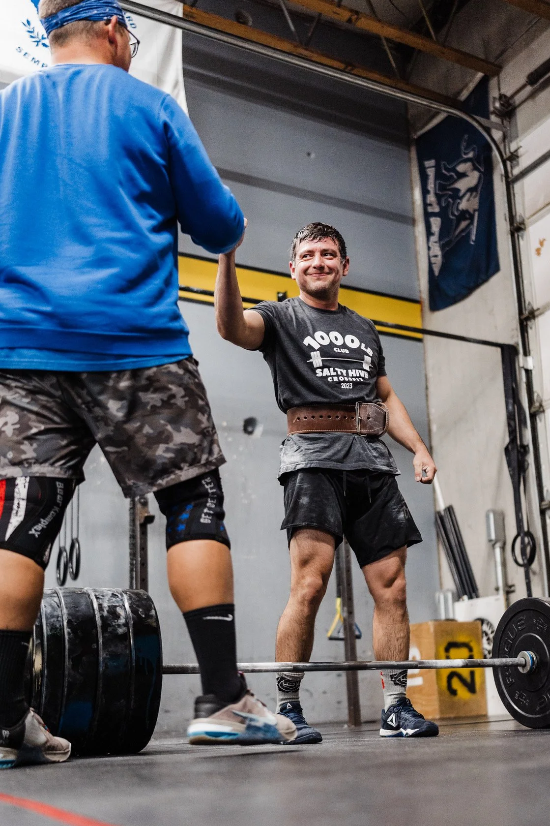 Two men in a gym, one lifting weights and the other congratulating him with a handshake. The man lifting weights is wearing a black shirt with white text, camouflage shorts, knee sleeves, and weightlifting shoes. The other man is wearing a blue hoodie, camouflage shorts, knee sleeves, and sneakers.