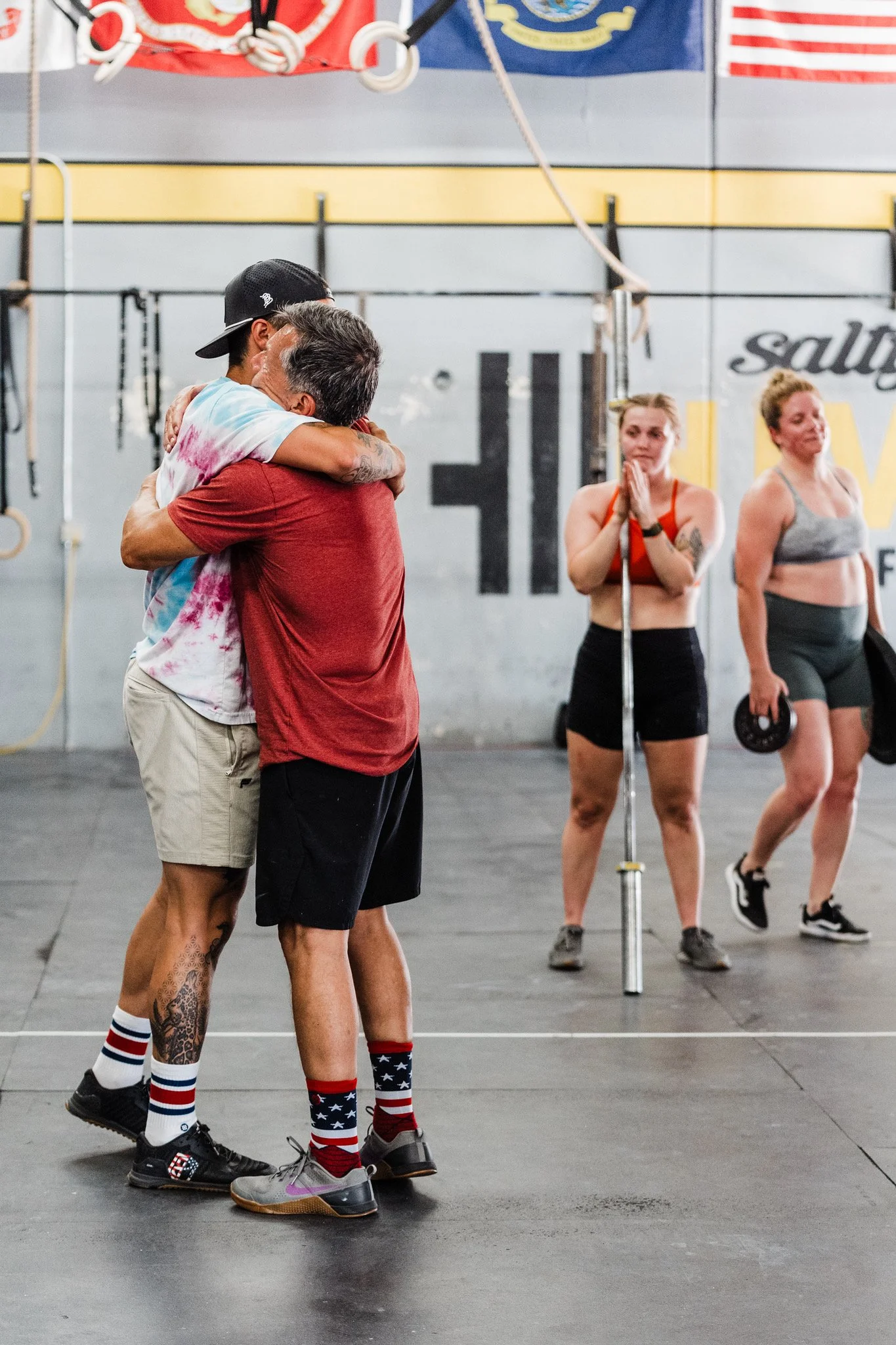 Two men hugging, one wearing a tie-dye shirt and the other wearing a red shirt, in a gym. Behind them are three women, one in a red sports bra, and two in gray sports bras, standing and observing. The gym has fitness equipment and flags hanging on the wall.