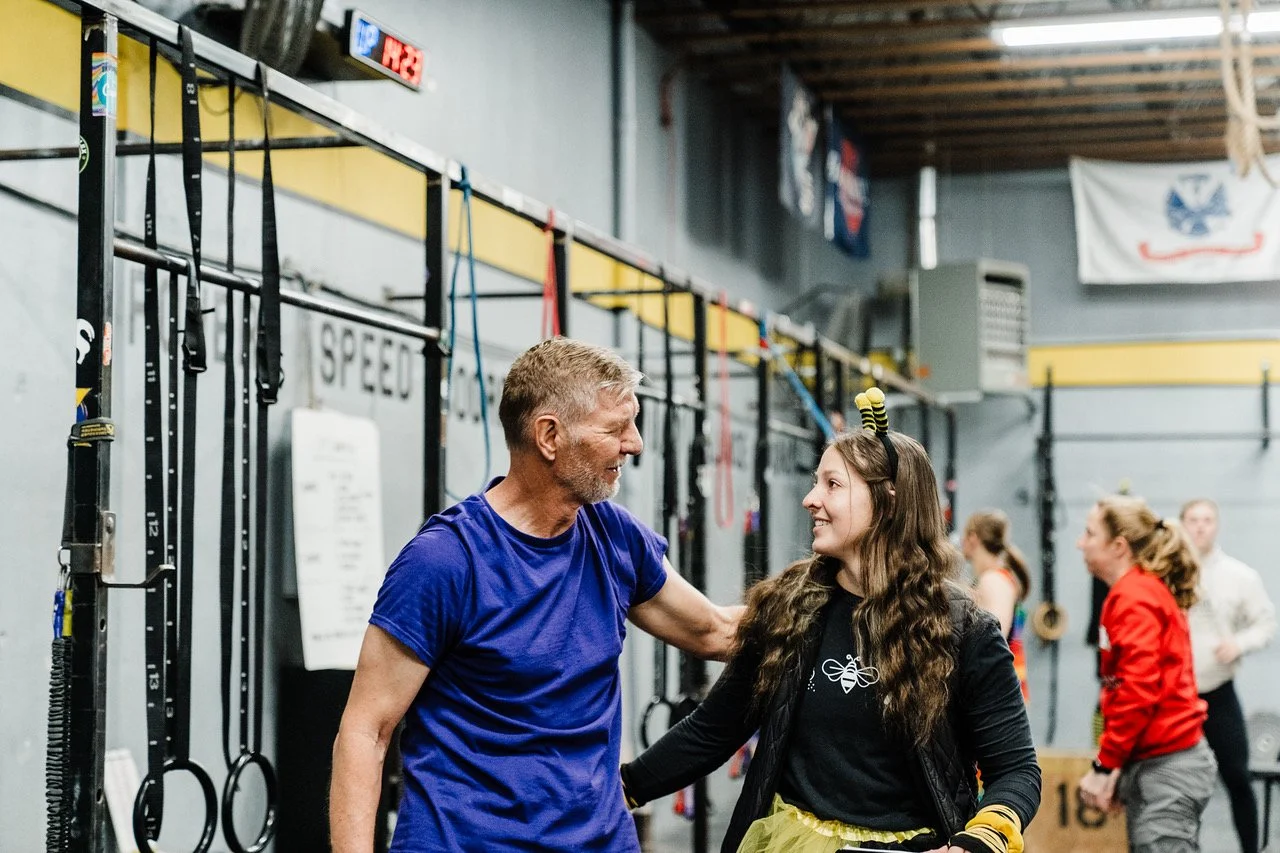 A man and a woman smiling at each other in a gym, with fitness equipment like rings in the background.