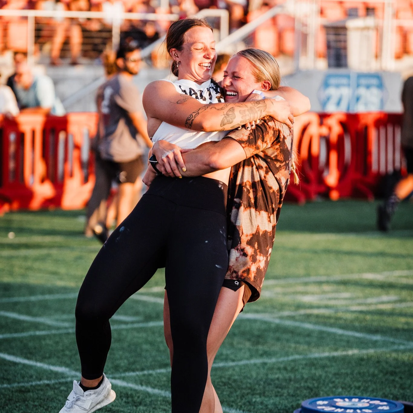 Two women hugging on a sports field, smiling and laughing, with people and orange barriers in the background.