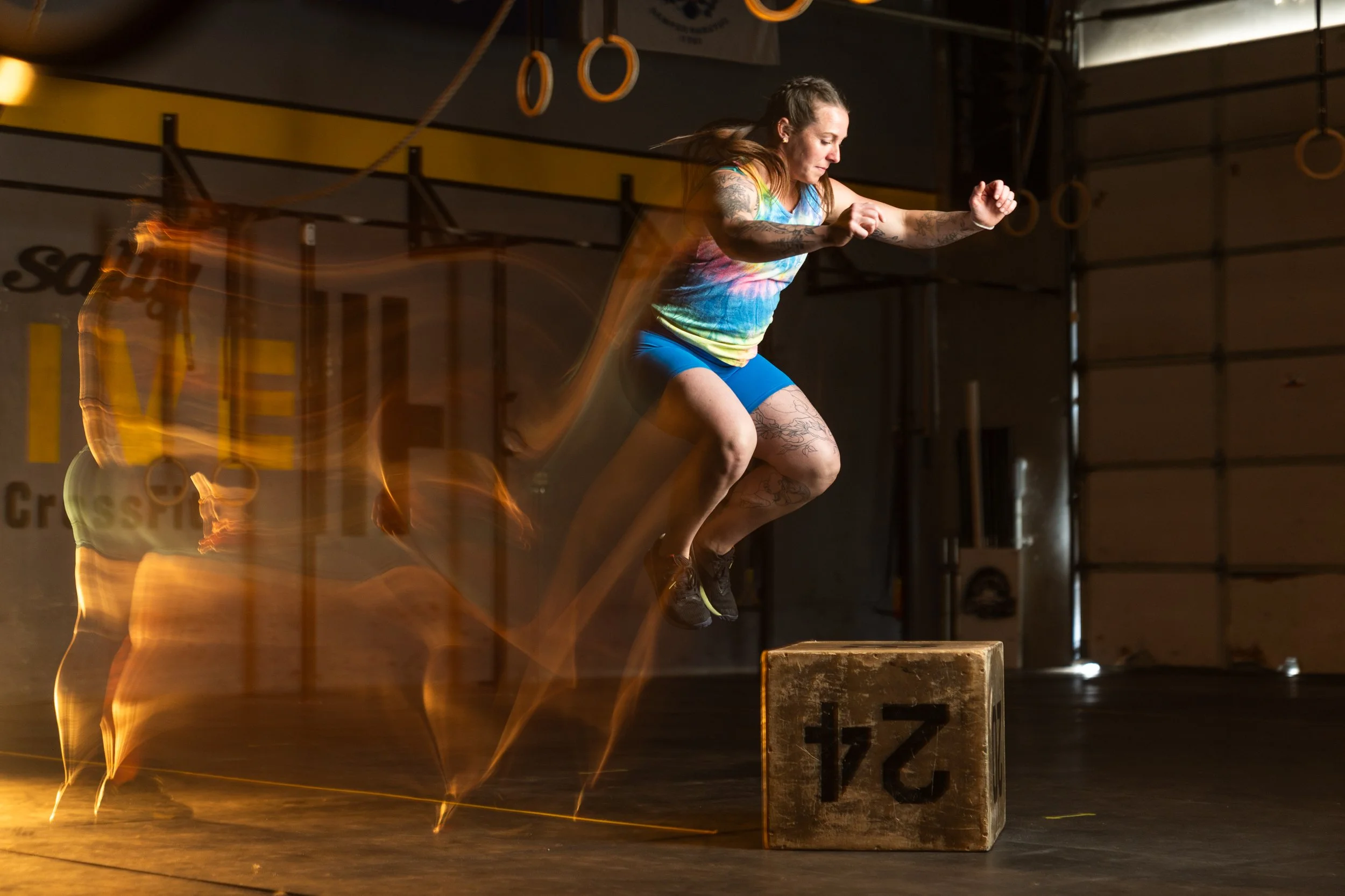 A woman in workout clothes is mid-air performing a jump onto a wooden box in a gym. The background shows gym equipment and gymnastic rings hanging from the ceiling.