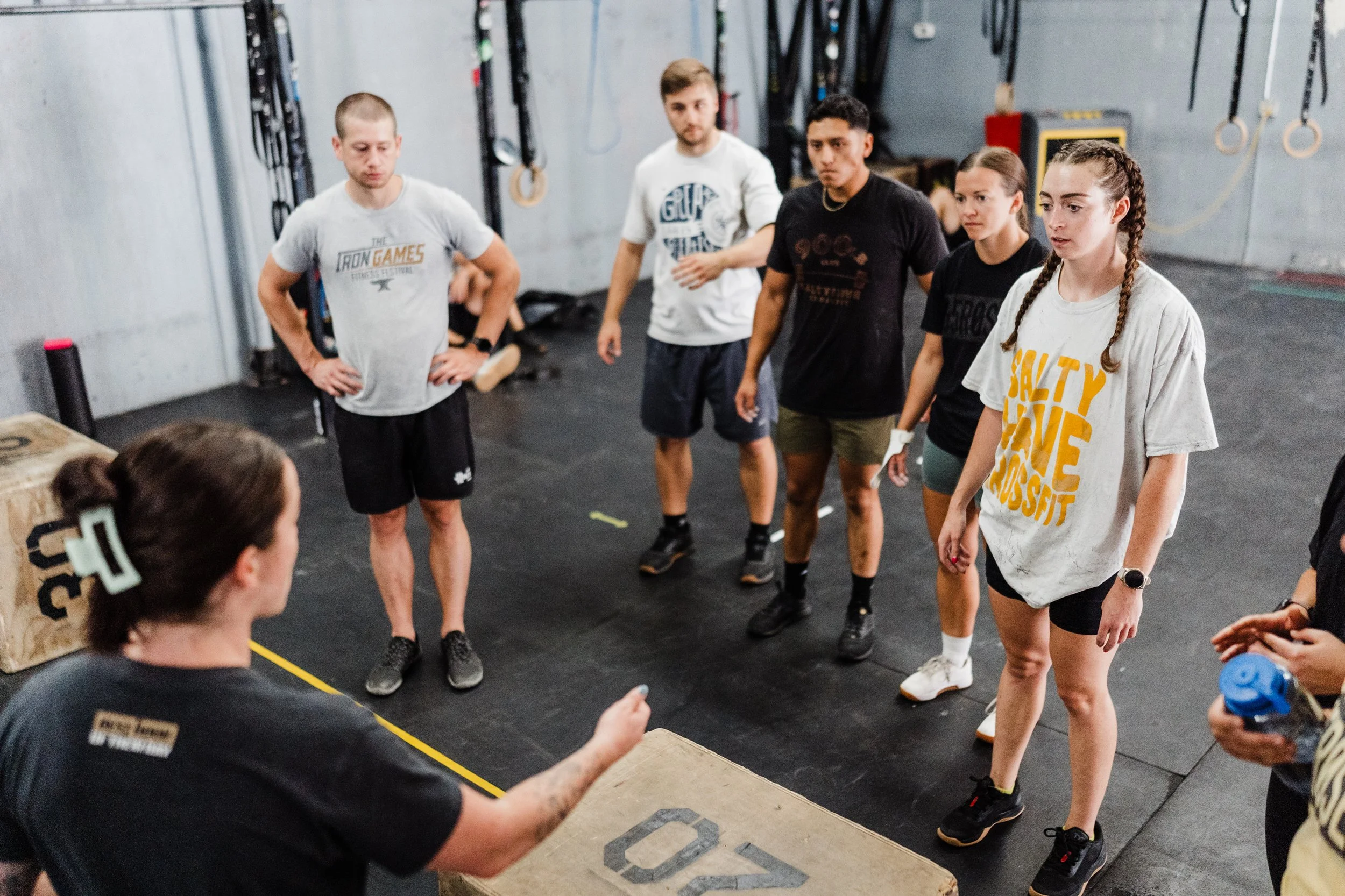 A group of five people standing in a gym, listening to a woman who appears to be instructing or coaching them. The gym has fitness equipment hanging on the walls and gymnastic rings. The people are wearing workout clothes and seem attentive.