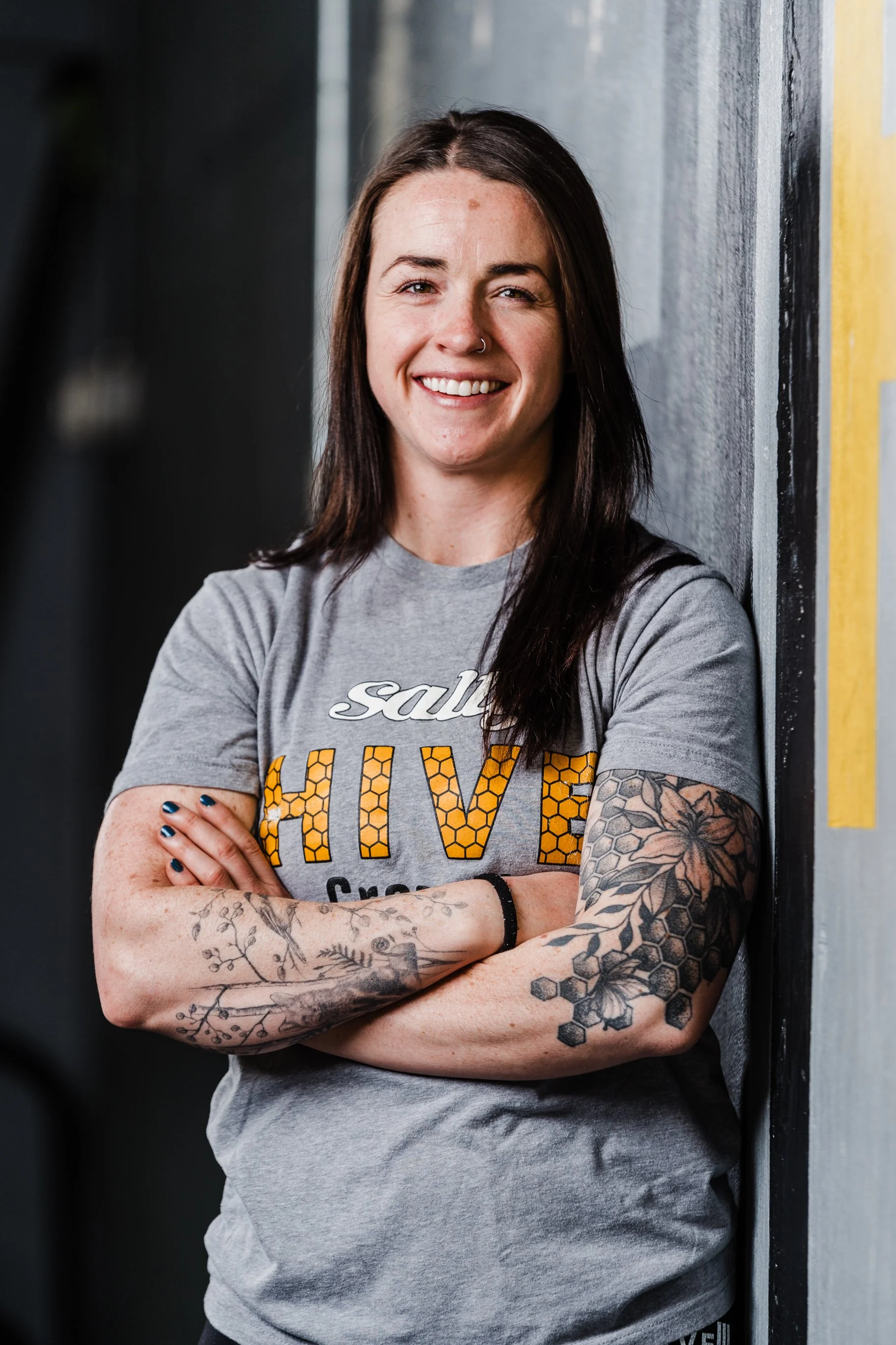A woman with dark brown hair, tattoos on her arms, and a nose ring, smiling and leaning against a wall with her arms crossed, wearing a gray t-shirt.