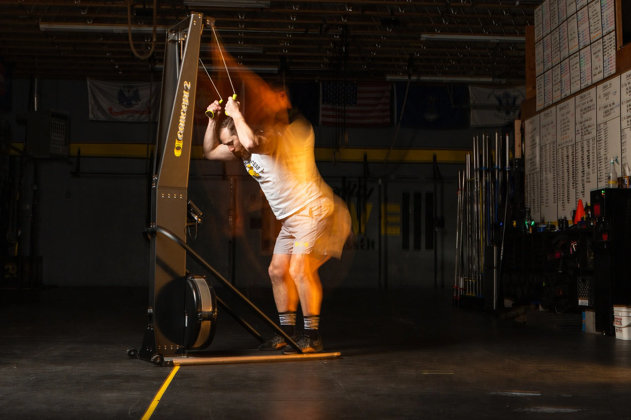 A man in a gym operates a skiing machine while stretching his arm over his head. The gym has various equipment and a large menu or schedule board on the wall in the background.