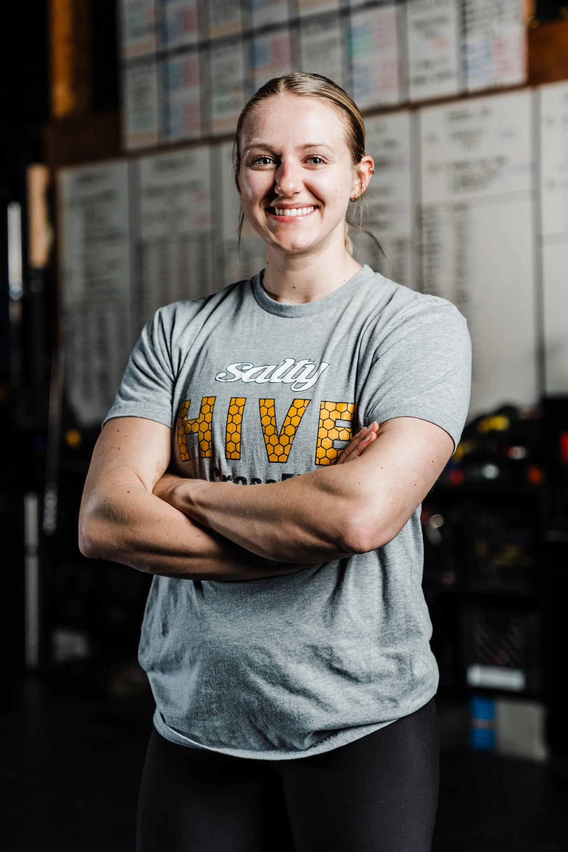Young woman smiling with arms crossed, wearing a gray T-shirt with 'salty' and 'HIVE' printed on it, in a gym setting.