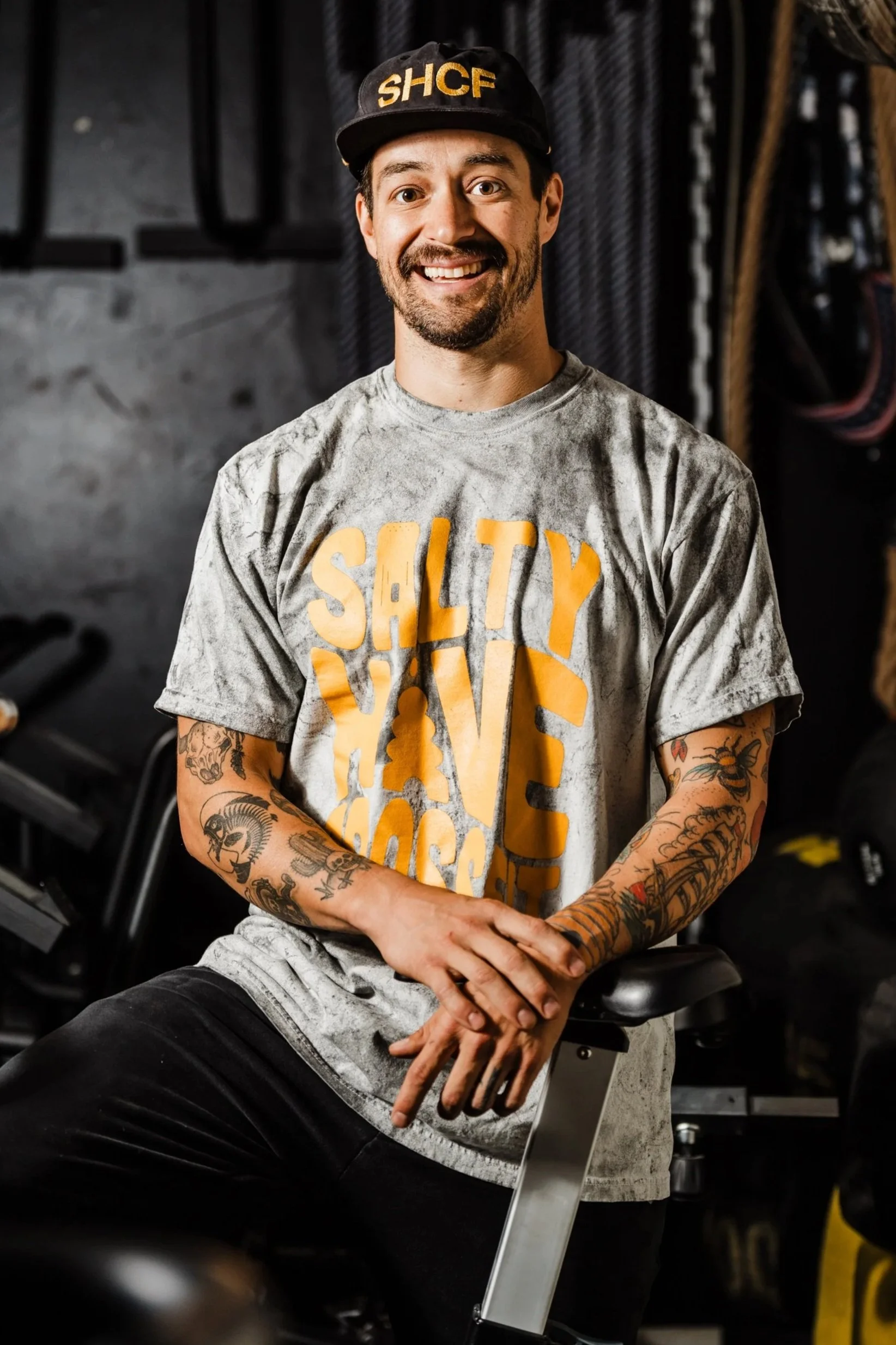 A man with tattoos on his arms, wearing a gray T-shirt with large yellow text and a black cap with yellow text, sitting in a gym or fitness studio with black exercise equipment in the background.