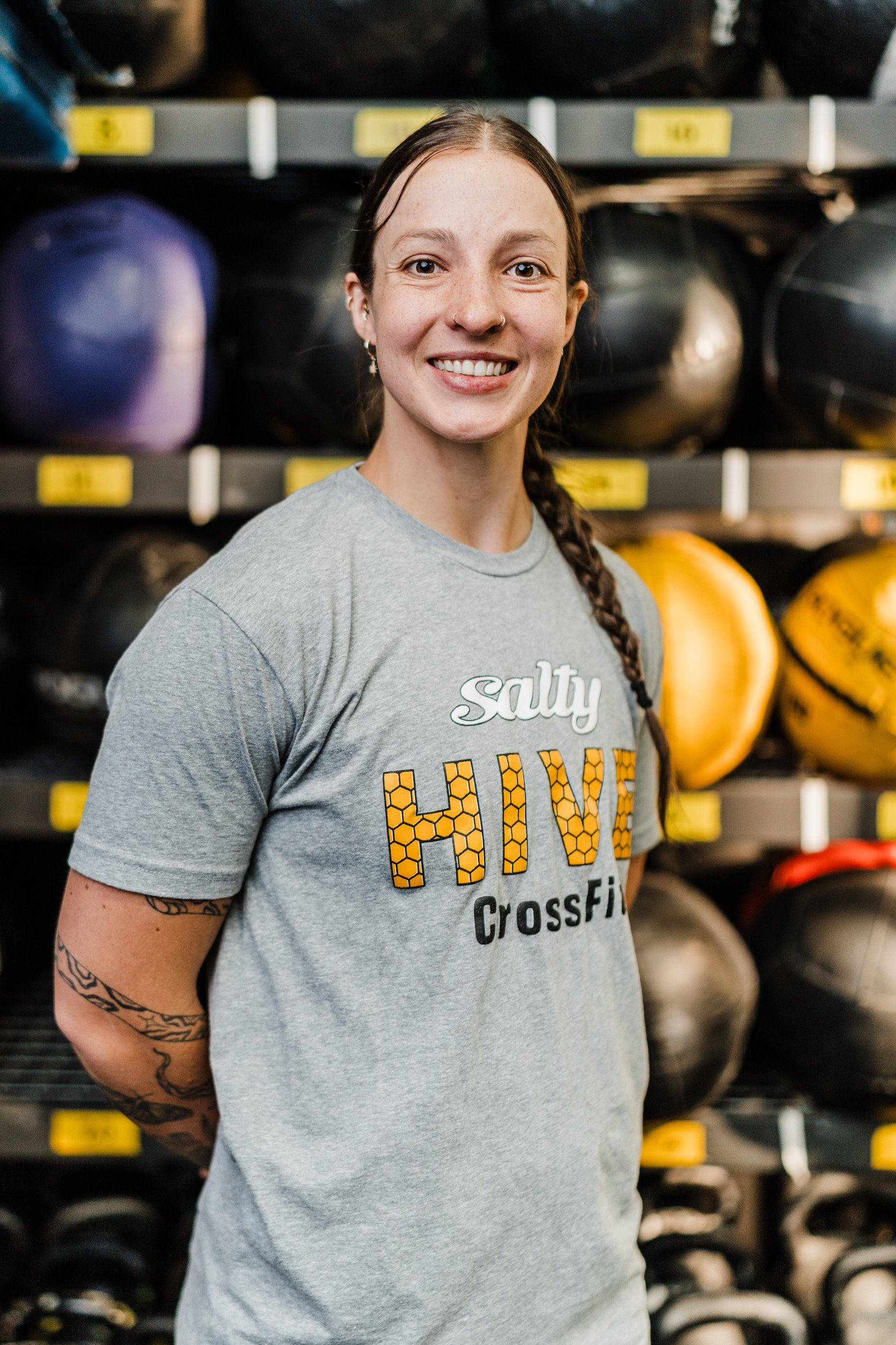A young woman with brown hair in a braid and tattoos on her arm, smiling, wearing a gray CrossFit t-shirt, standing in front of shelves filled with medicine balls.