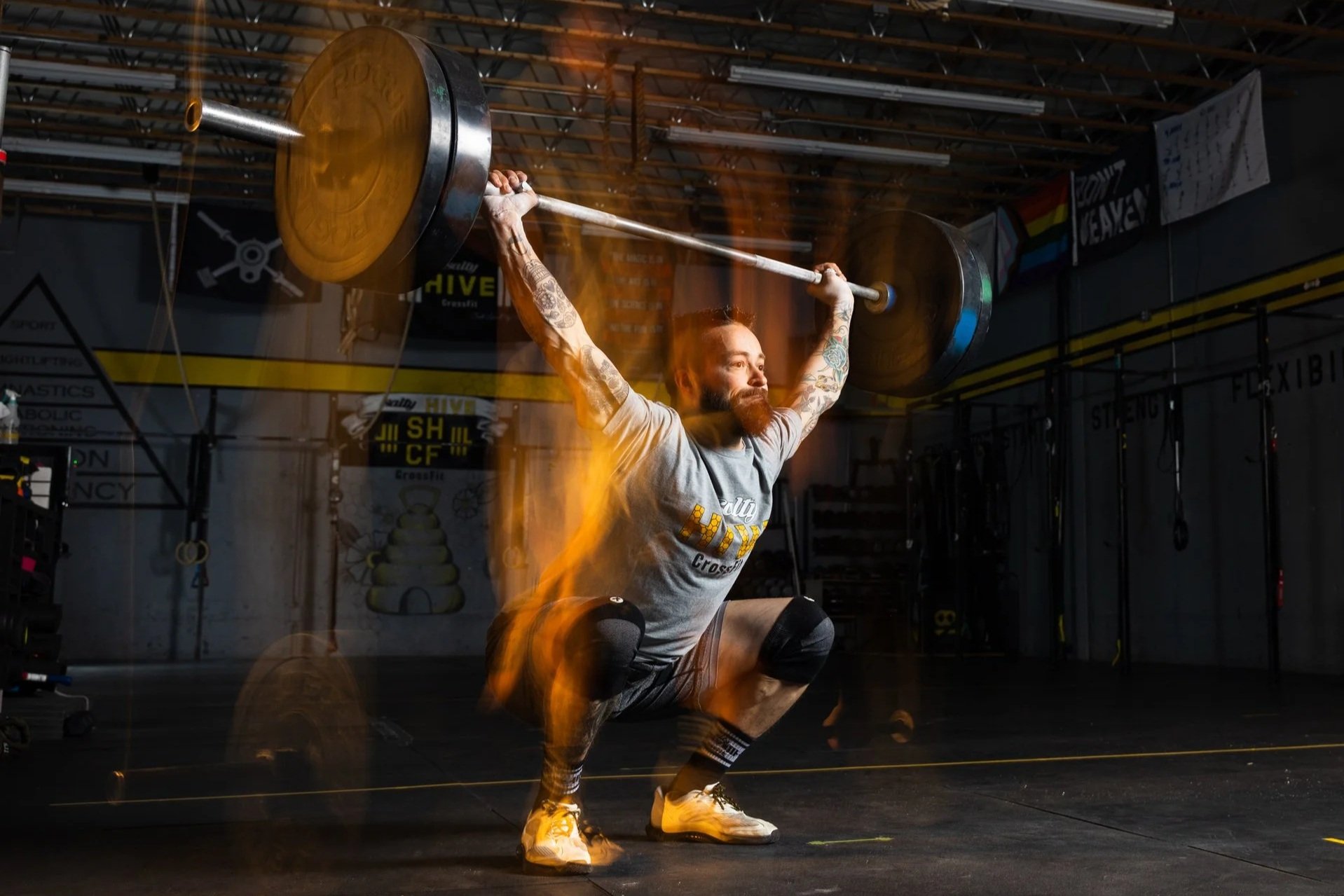 A man with tattoos and a beard performing an overhead squat with a barbell in a gym. Flames and sparks surround him, and the gym has various equipment and banners.