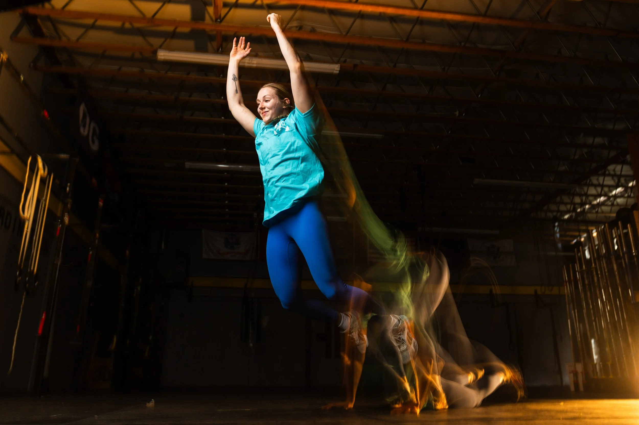 A woman in a blue shirt and blue leggings is jumping in the air with her arms raised inside a gym. The photo captures motion blur of her movement and the gym's dark interior, with some equipment visible in the background.
