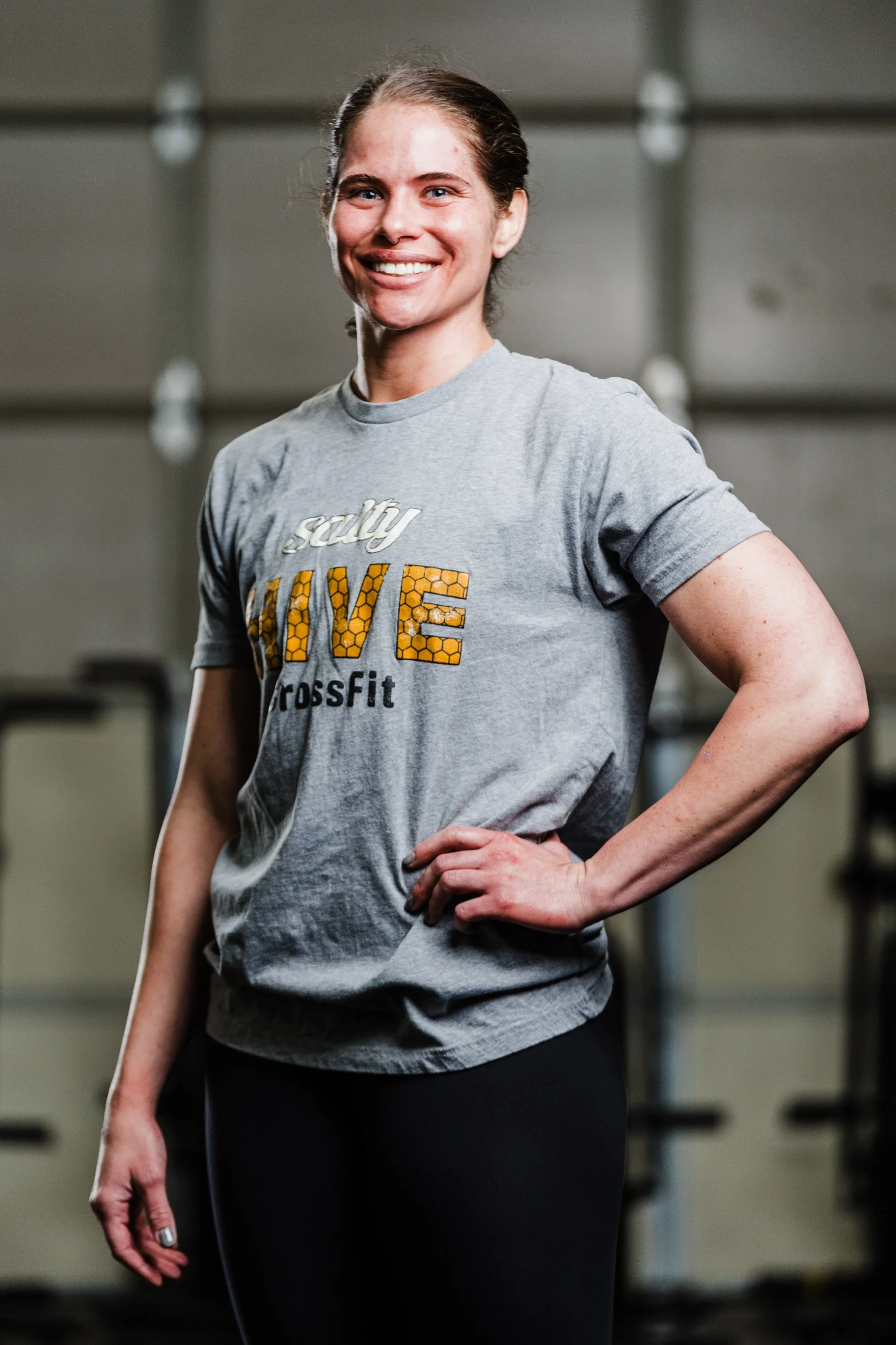 A smiling woman standing with her hand on her hip in a gym, wearing a gray t-shirt with the words "Salty HIVE CrossFit" and black leggings.