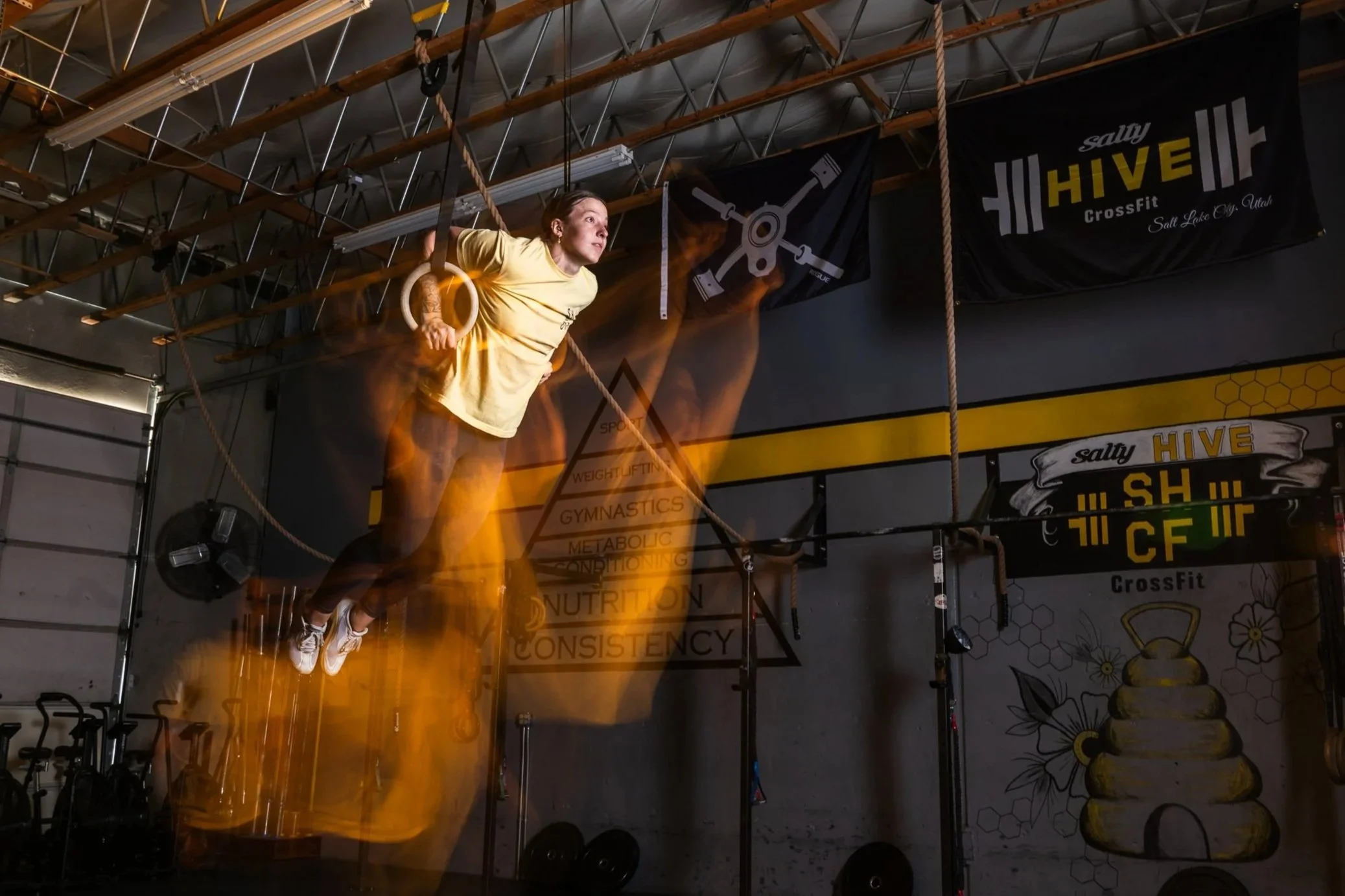 A woman performing an exercise on gymnastic rings in a fitness gym with CrossFit banners called 'Salty Hive'